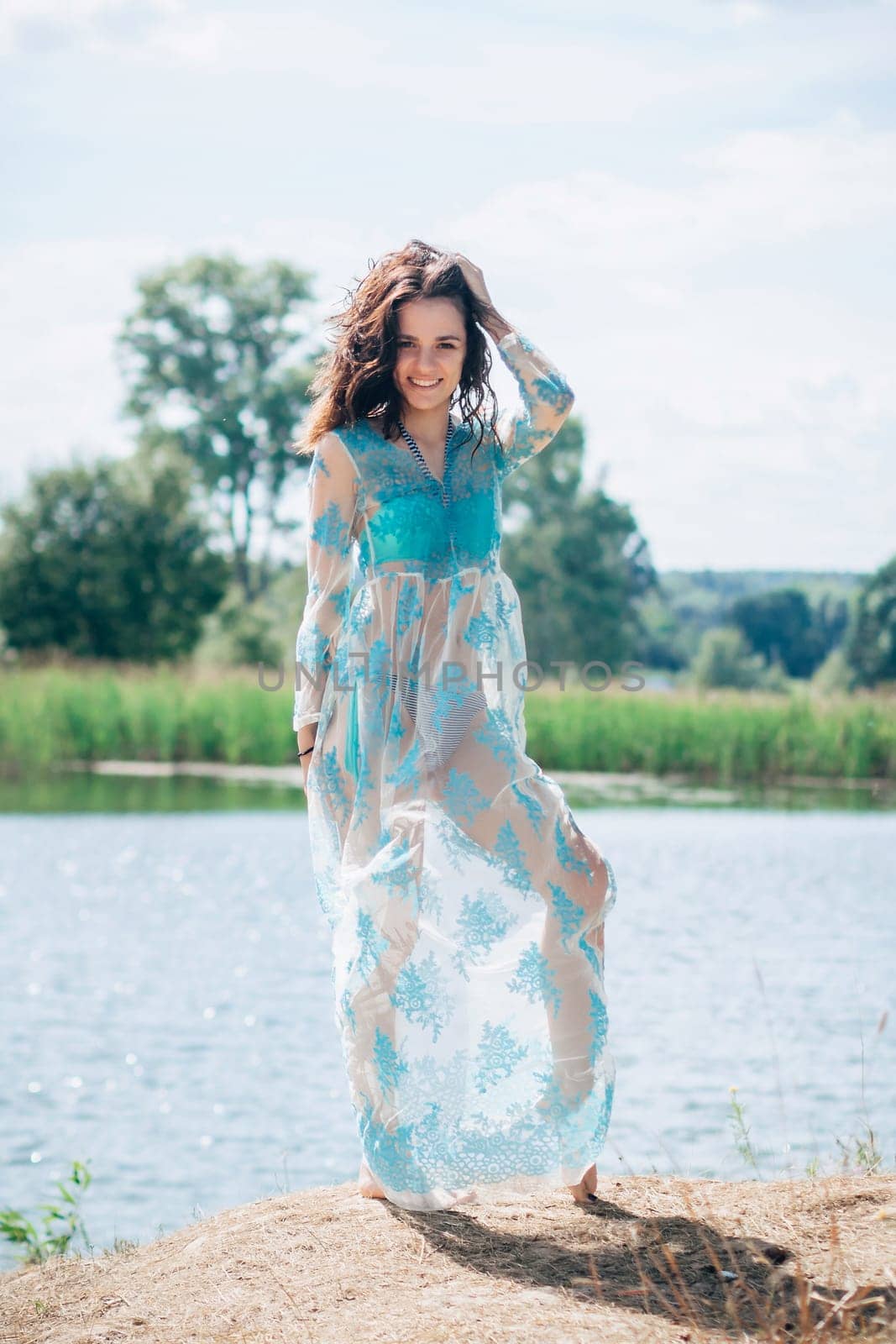 young girl with a sexy body in a swimsuit and tunic smiles on beach by lake in summer