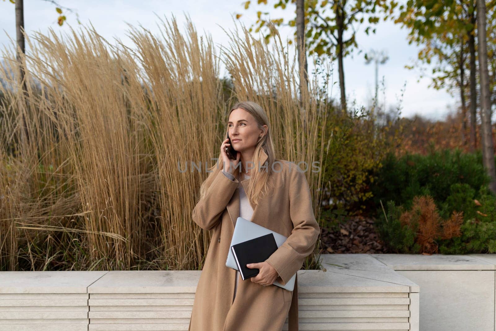 A woman wearing a tan coat stands near a building, her back to a wall, talking on the phone. She holds a laptop and a notebook. Tall grass grows in the background.