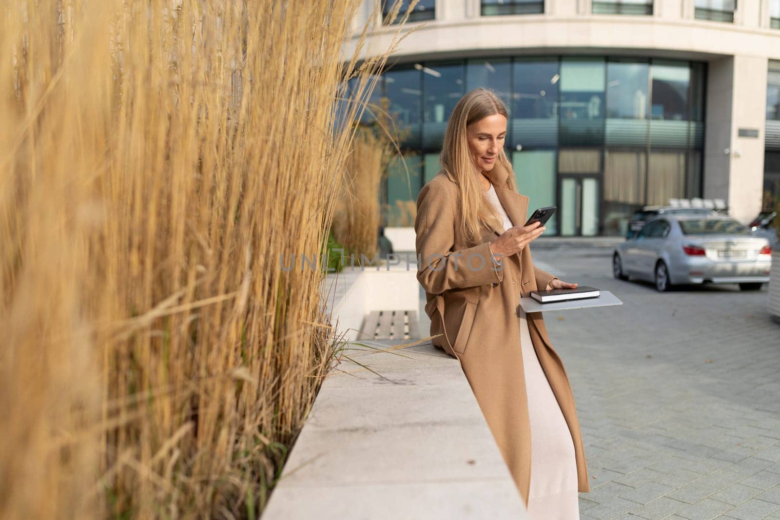 A businesswoman with long blonde hair is wearing a beige coat. She is standing outside a business center and using her phone. She has a notebook in her other hand.