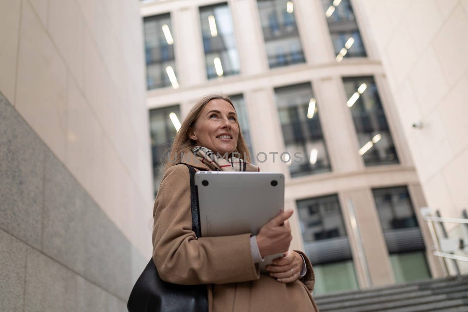 A professional woman is walking near a sleek business center. She carries a laptop and smiles, embodying confidence in a bustling urban environment.