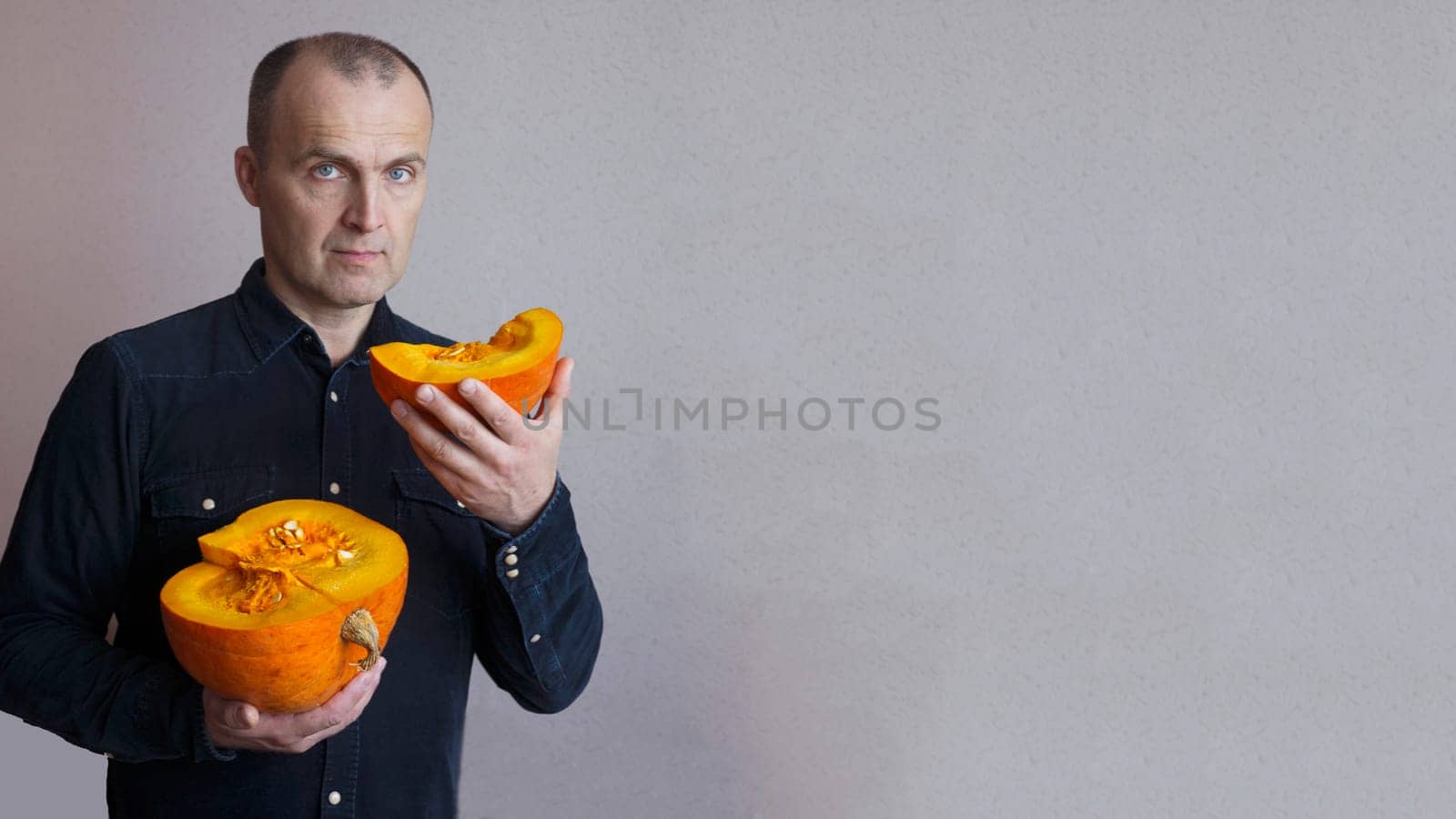 A handsome elderly man in a blue shirt against the wall holds a cut orange pumpkin with his hands and looks at the camera.