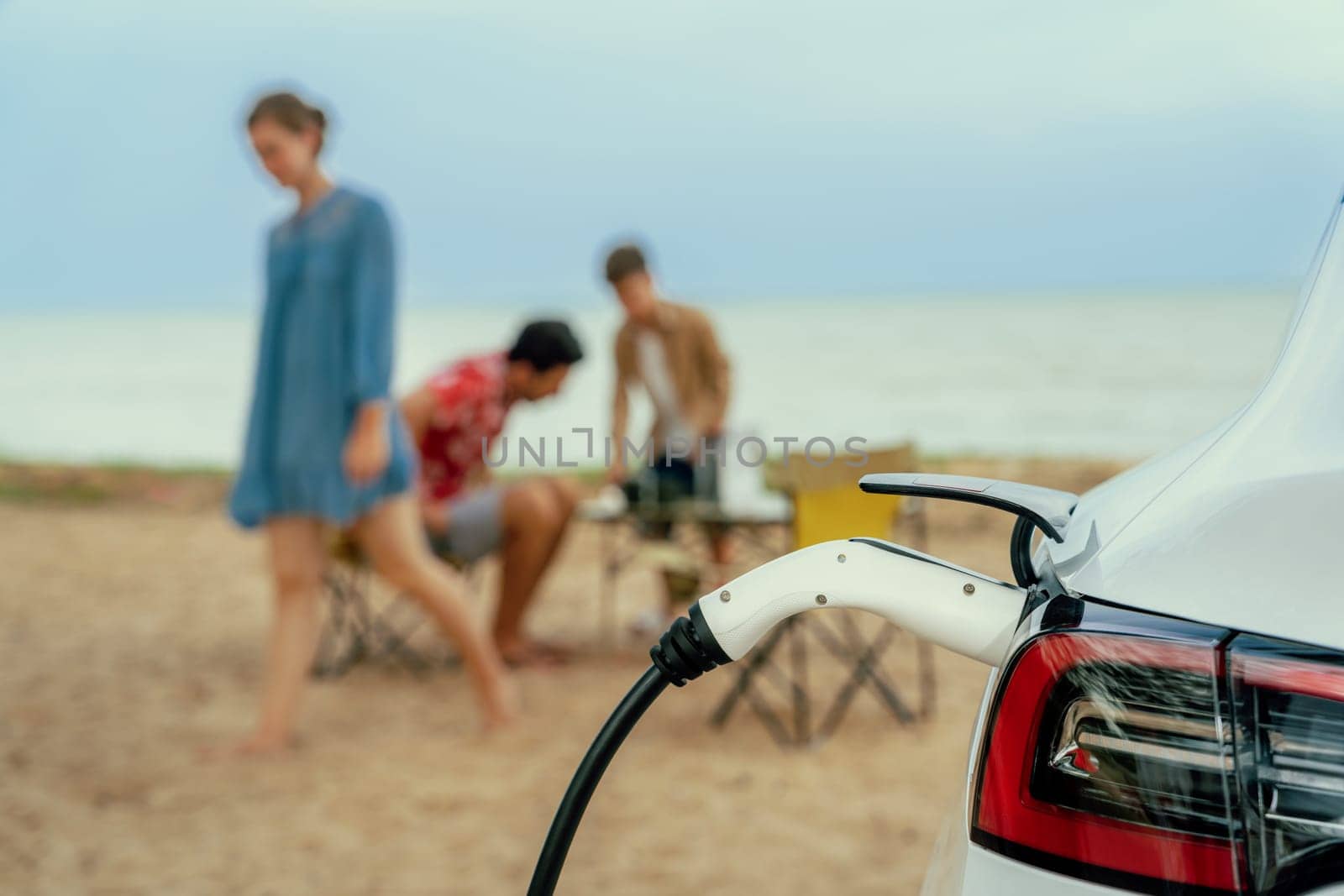 Alternative family vacation trip traveling by the beach with electric car recharging battery from EV charging station with blurred family enjoying the seascape background. Perpetual