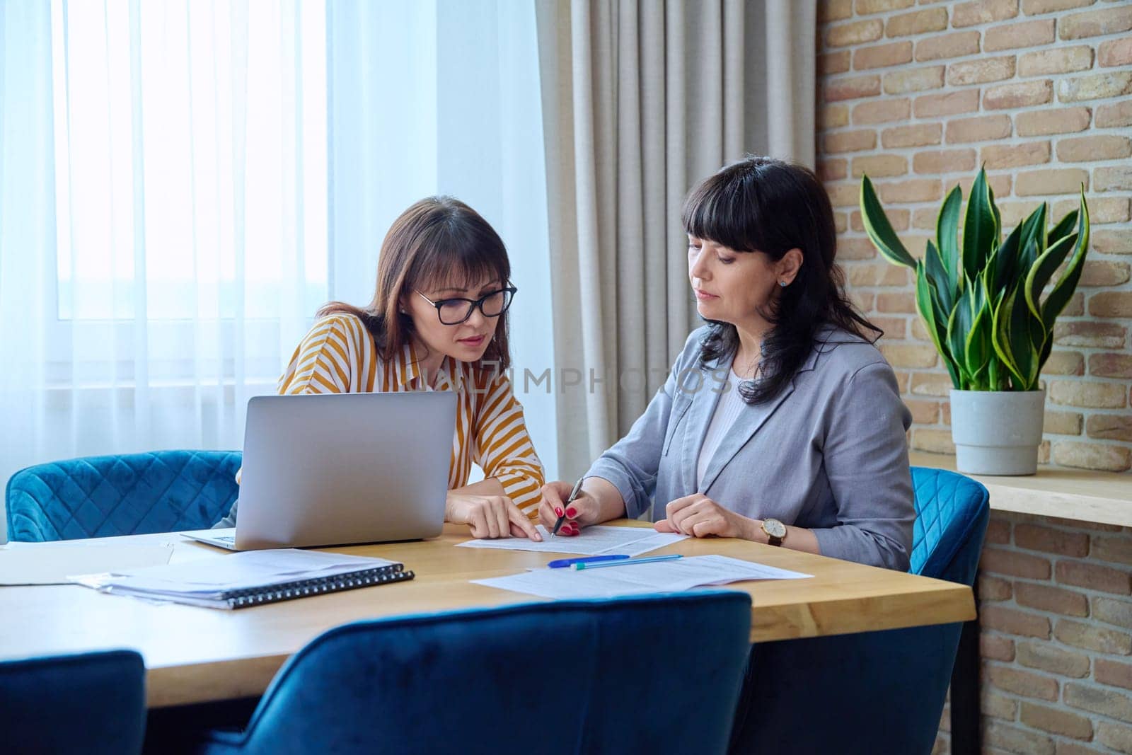 Two mature business women colleagues sitting at large table in office with business papers contracts laptop computer. Business ceo work law finance mentoring consulting teamwork, 40s 50s people on job