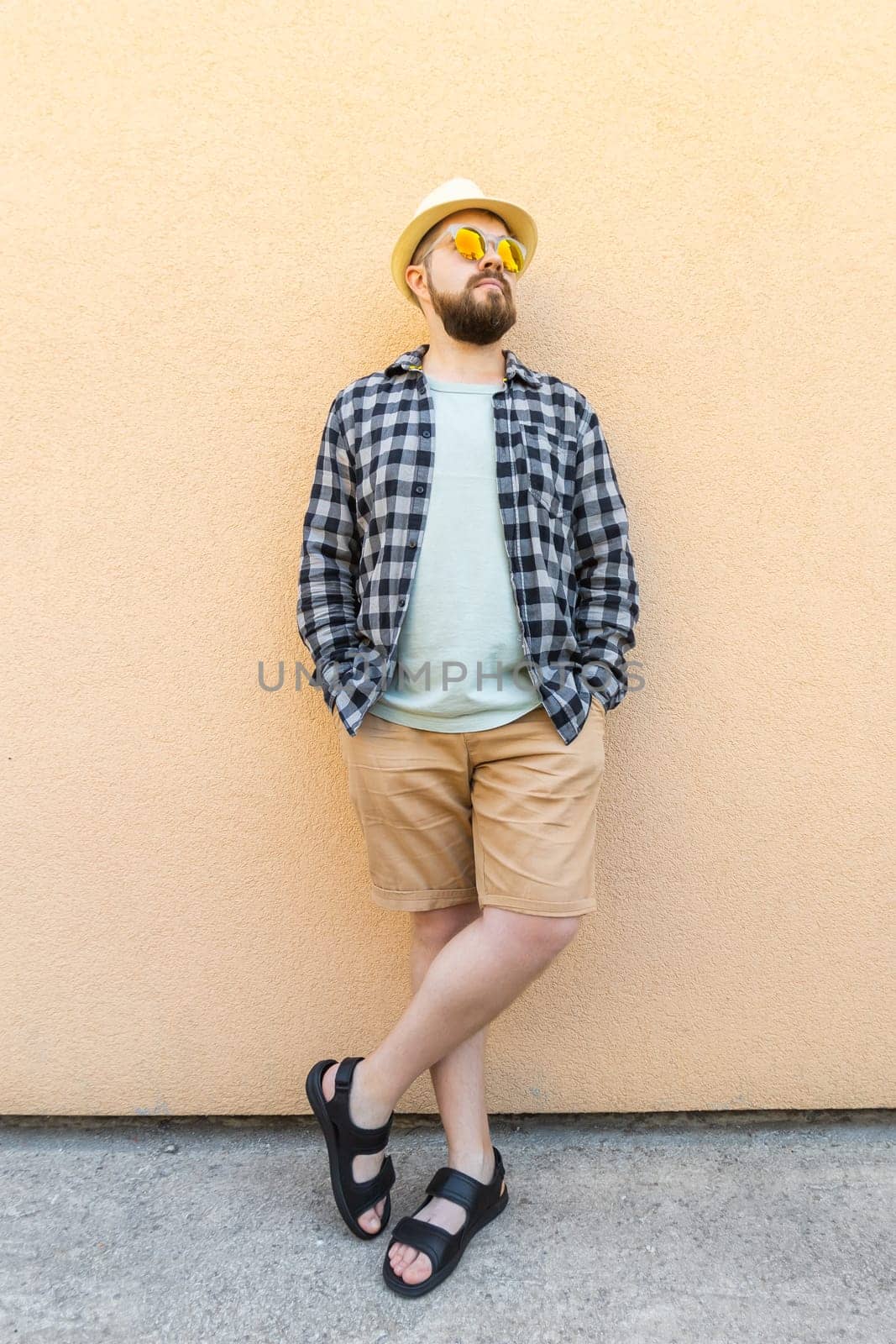 Bearded millennial man in summer clothes and hat stand over beige wall on street city urban and travel