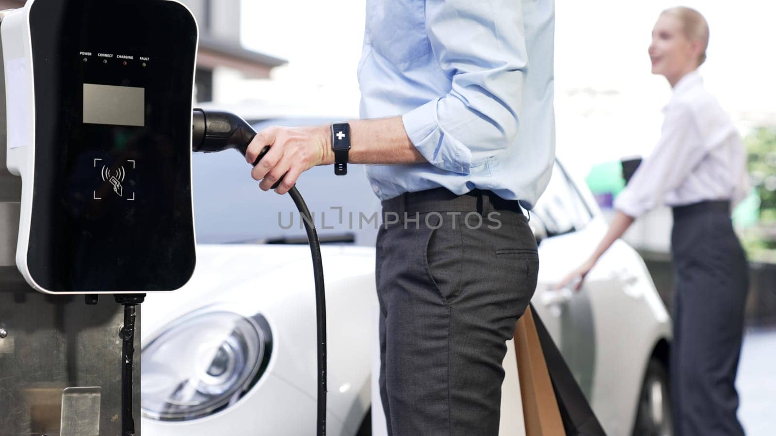 Businessman and businesswoman with progressive ambition unplug power cable from electric vehicle, standing on charging station with power cable plug and renewable energy-powered electric vehicle.