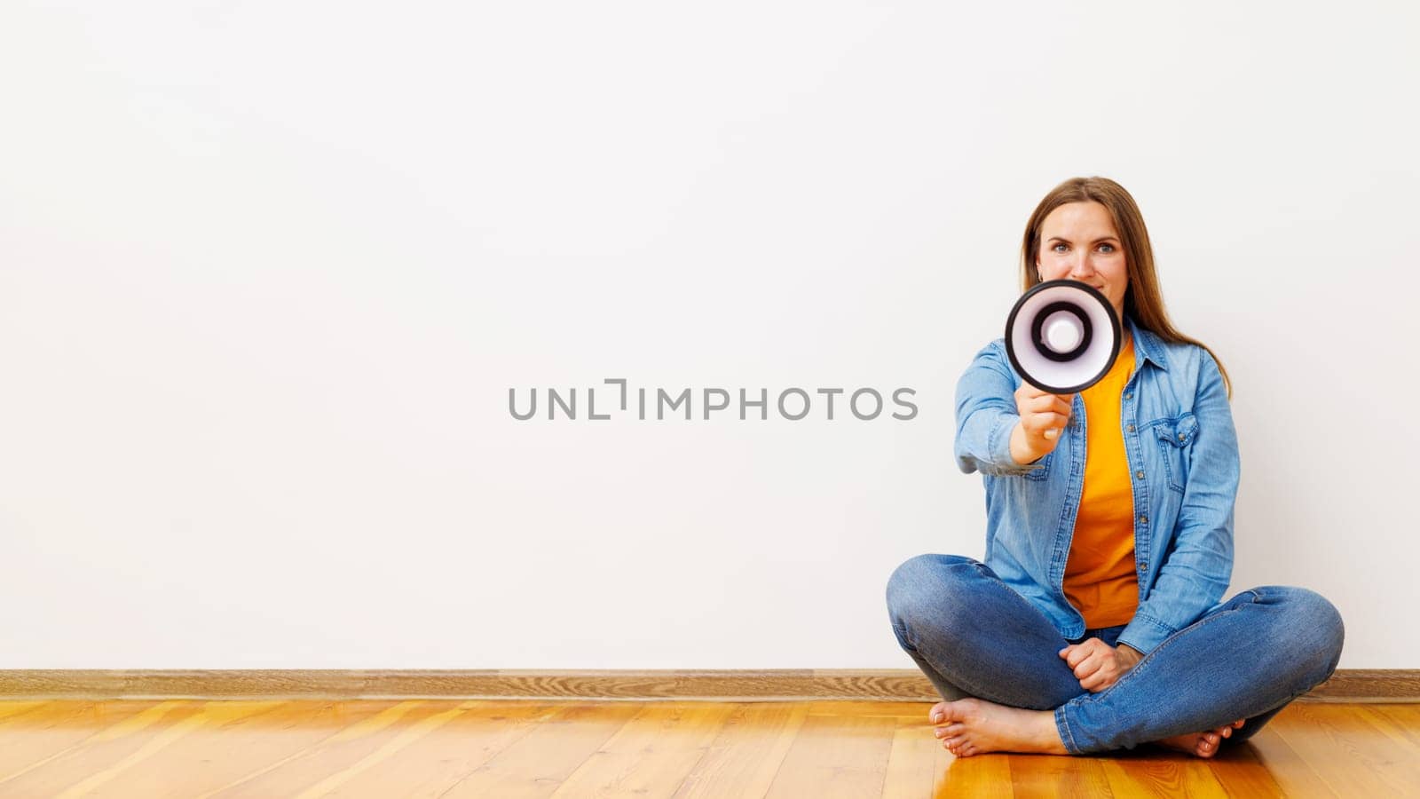 Young beautiful woman screaming on a megaphone sitting on a wooden floor in front of white wall.