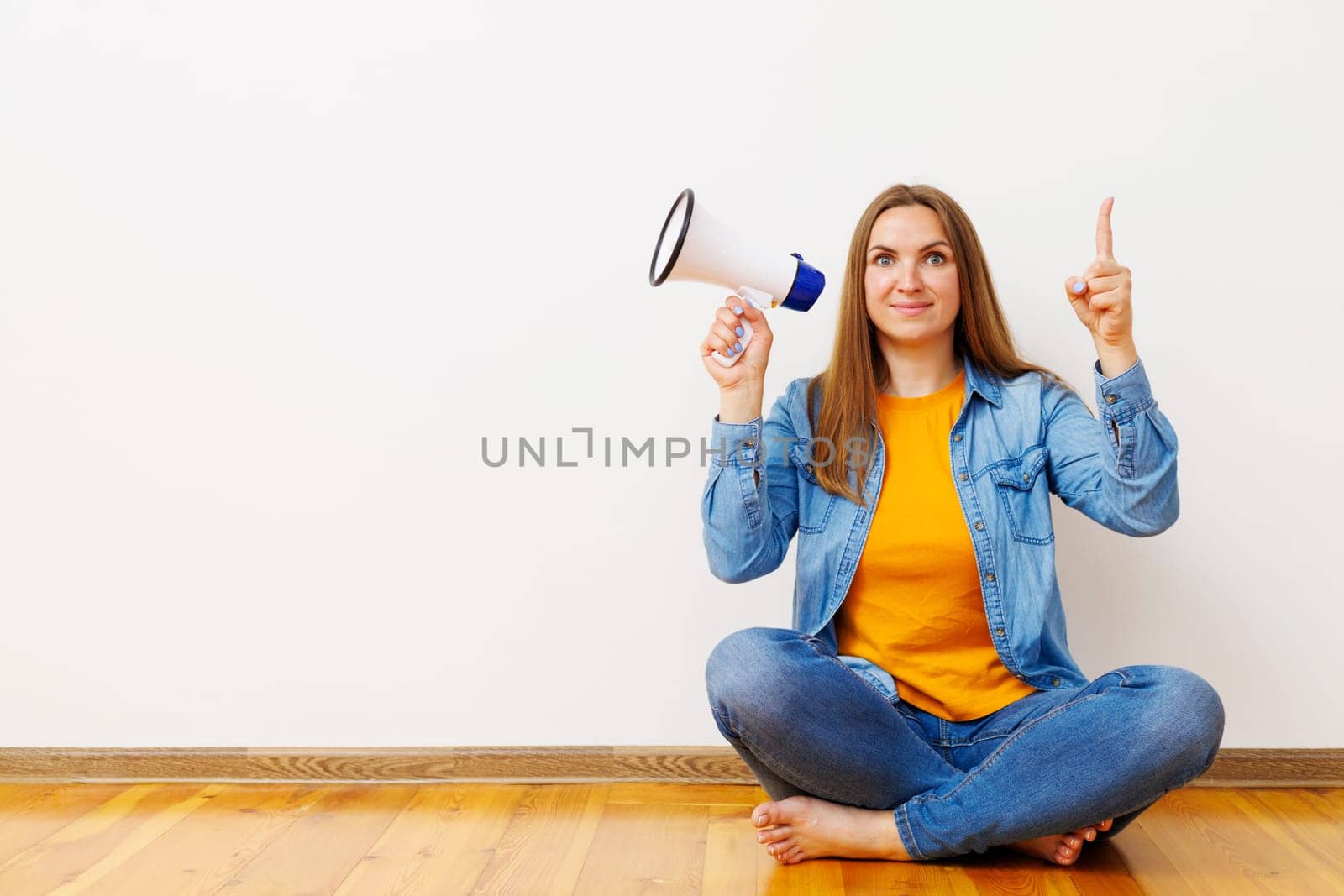 Young beautiful woman with megaphone sitting on wooden floor in front of white wall, copy space.