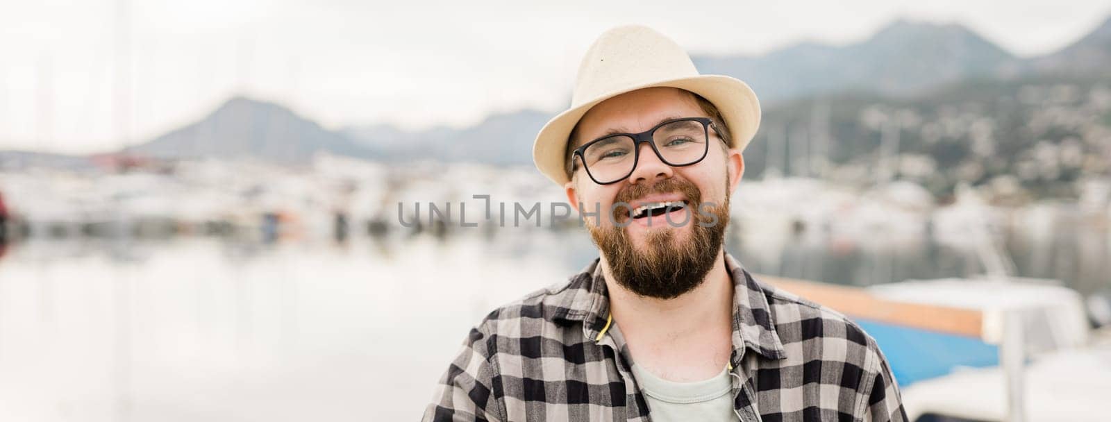 Banner millennial man wearing hat and glasses near marina with yachts. Portrait of laughing man with sea port background with copy space.