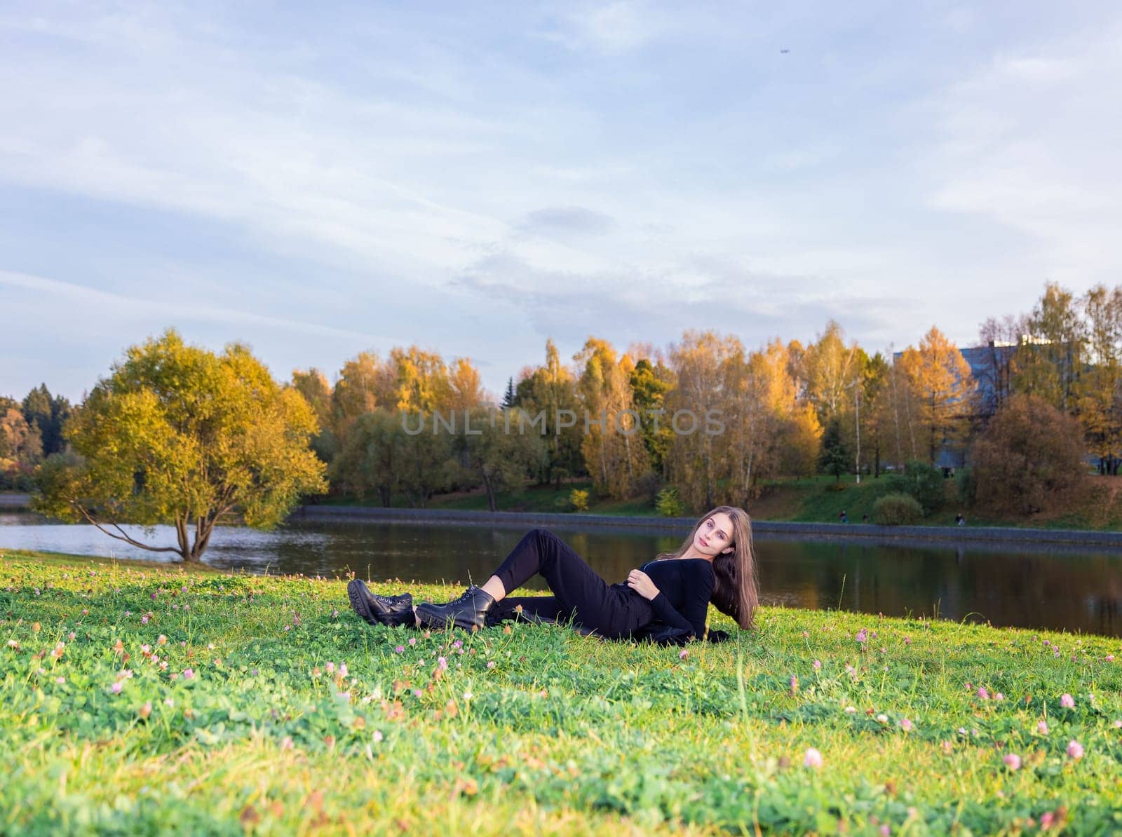 A beautiful girl poses while sitting on the grass by a pond in an autumn park