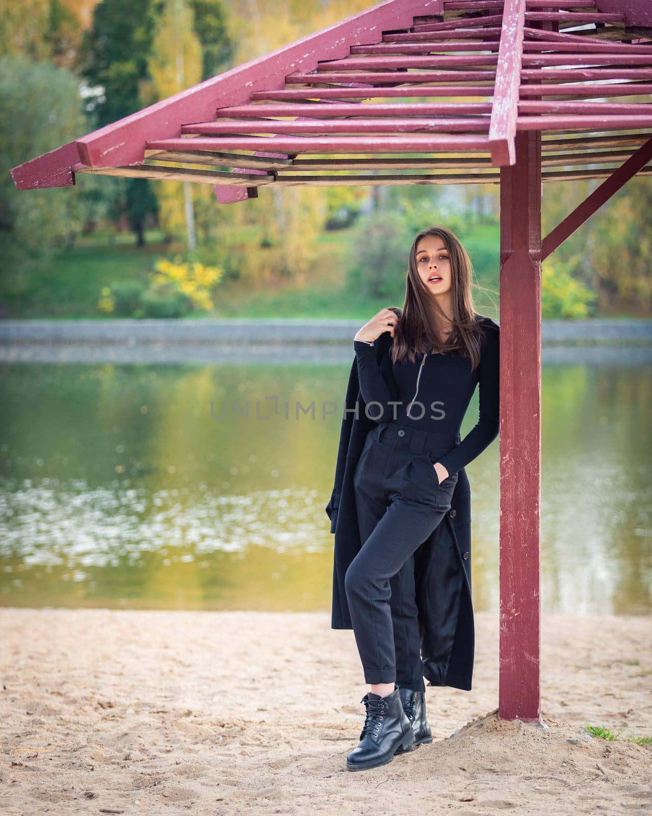 A beautiful girl poses while standing by a pond under an umbrella in an autumn park