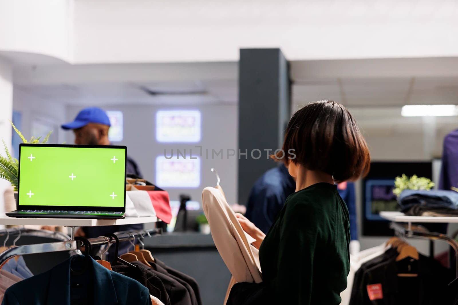 Open laptop with blank green screen in shopping mall, computer pc with mock up chroma key display standing on garment rack in fashion boutique. Woman shopper buying clothes during sales