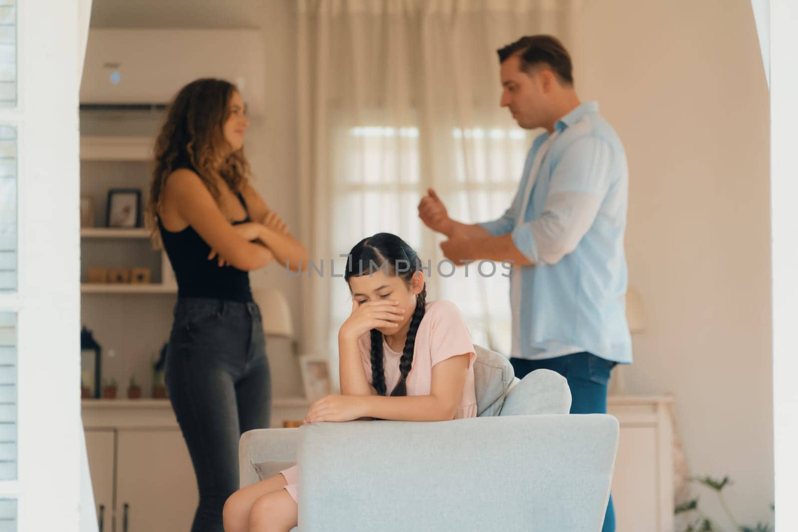 Annoyed and unhappy young girl sitting on sofa trapped in middle of tension by her parent argument in living room. Unhealthy domestic lifestyle and traumatic childhood develop to depression.Synchronos