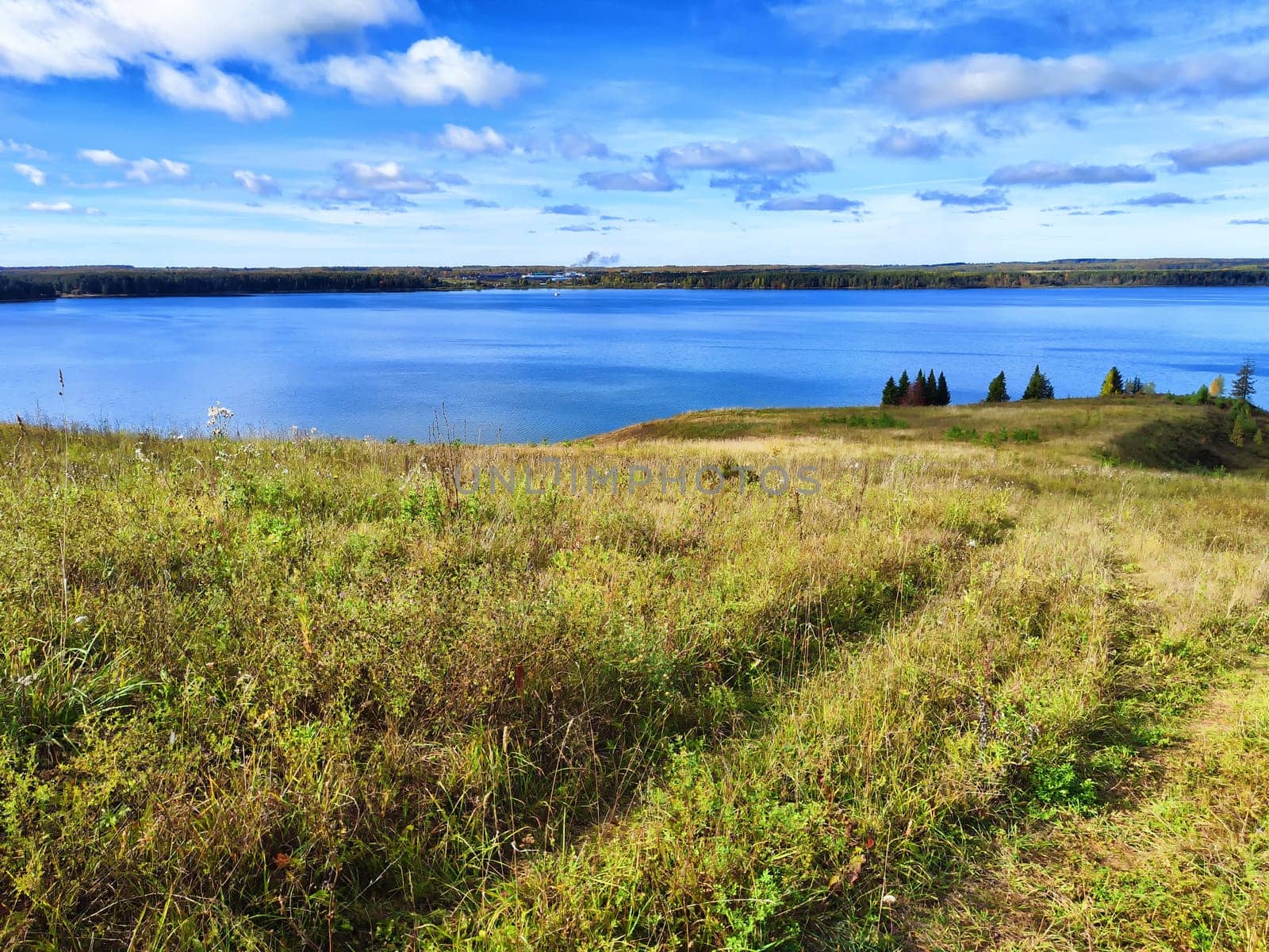 A serene lake stretches out beneath a blue sky with scattered clouds, framed by a grassy hillside.