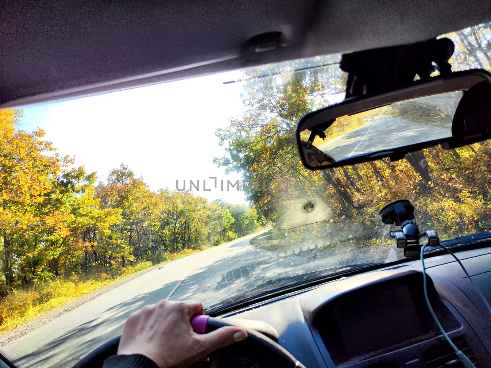 A person grips the steering wheel while driving down peaceful highway lined with autumn foliage and bright sunshine