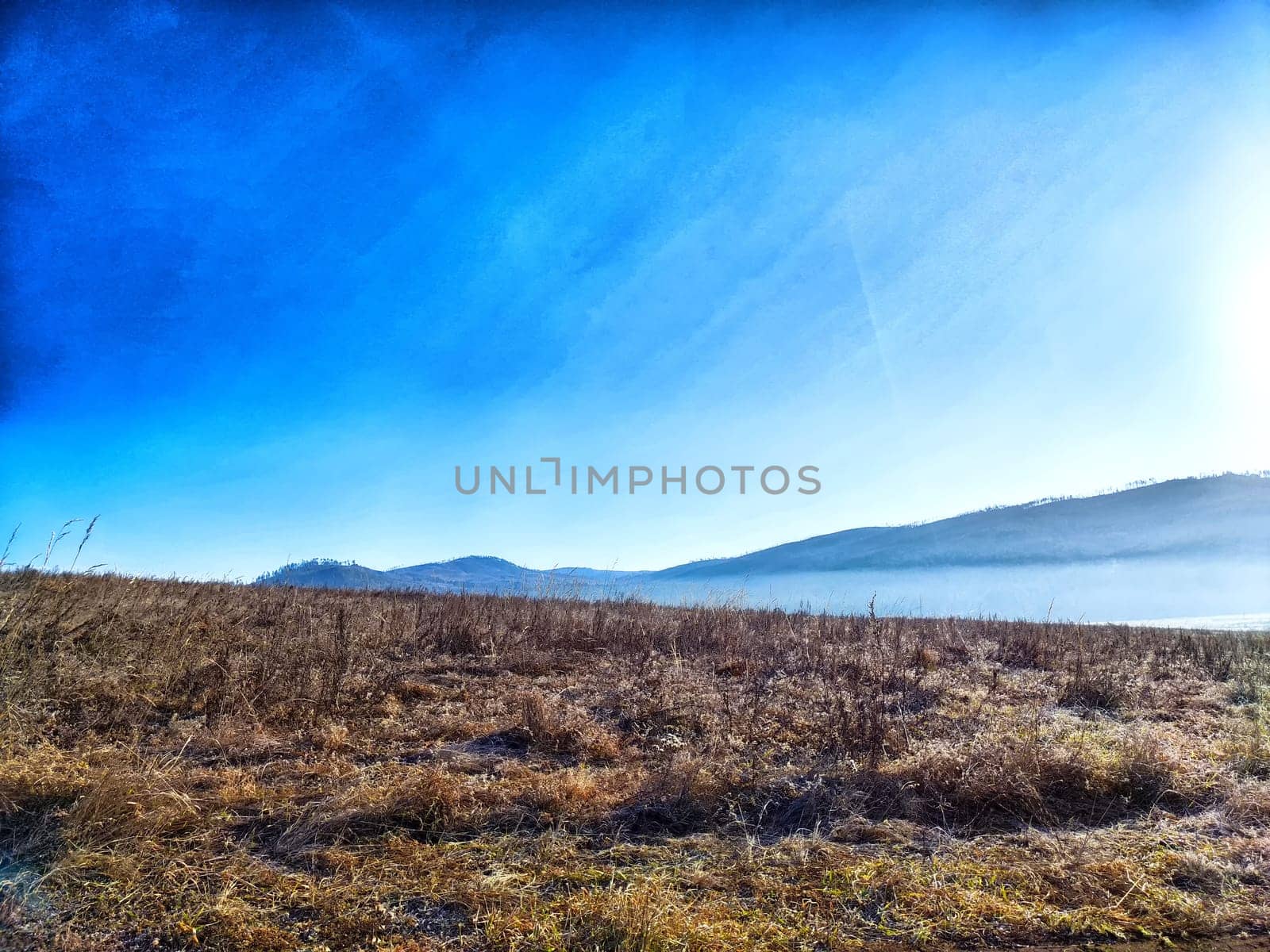 The landscape features a tranquil scene of a grassy field under a bright blue sky with mist settling over distant mountains.