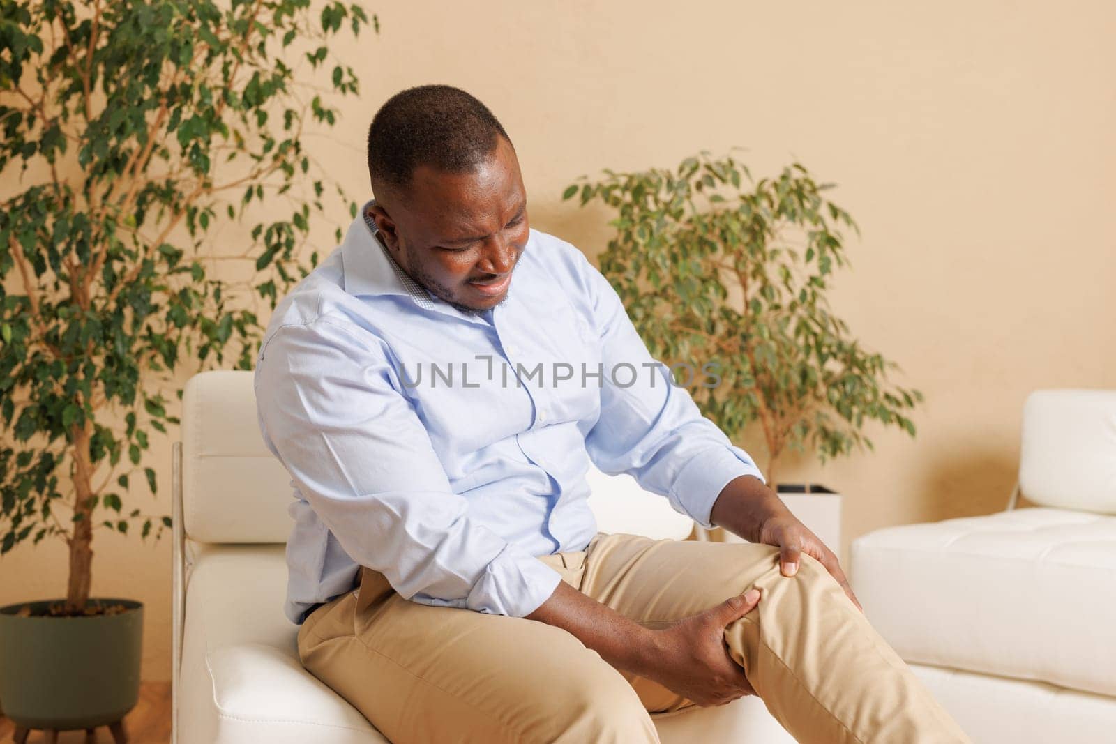 Knee pain sick body health care concept A young man has knee pain, sitting on the sofa at home, touching his leg, feeling pain in his knee.