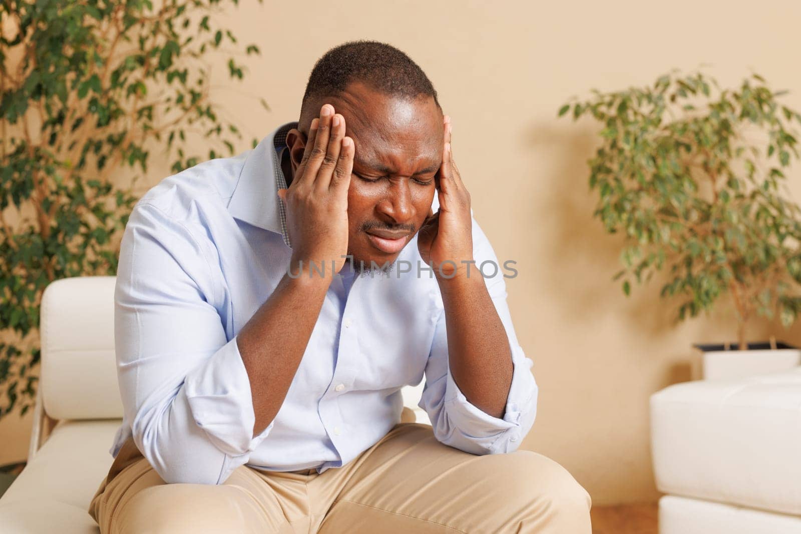 A man sitting on a couch, holding his head with both hands, showing signs of stress or headache. He is wearing a light blue shirt and beige pants, with a neutral background featuring plants.