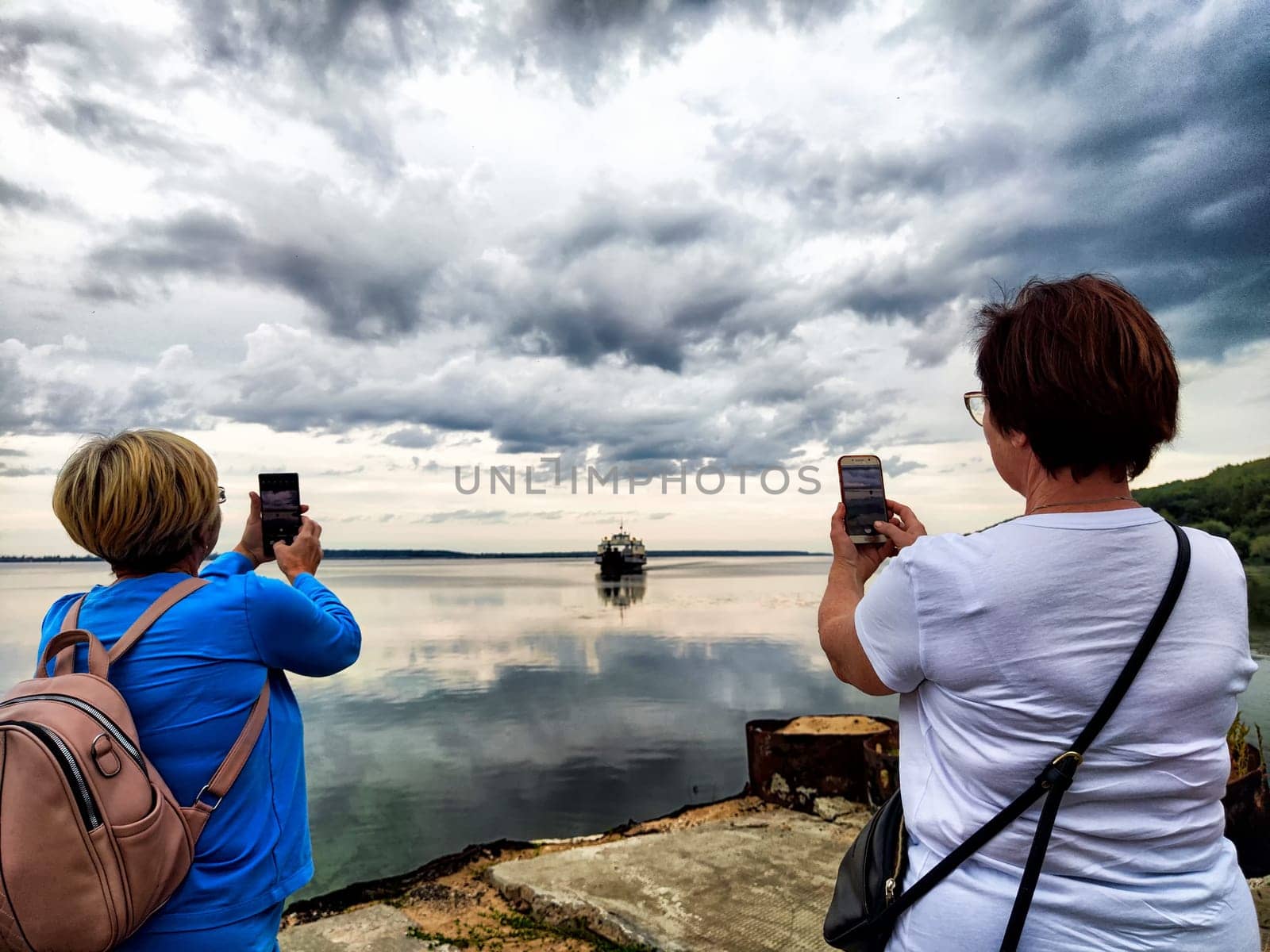 Standing by the lake, woman uses her phone to photograph the serene water and a boat against a backdrop of clouds