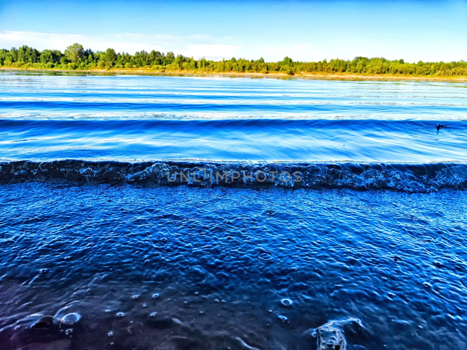 Gentle waves roll onto the rocky shore, reflecting the open blue sky and lush greenery nearby.