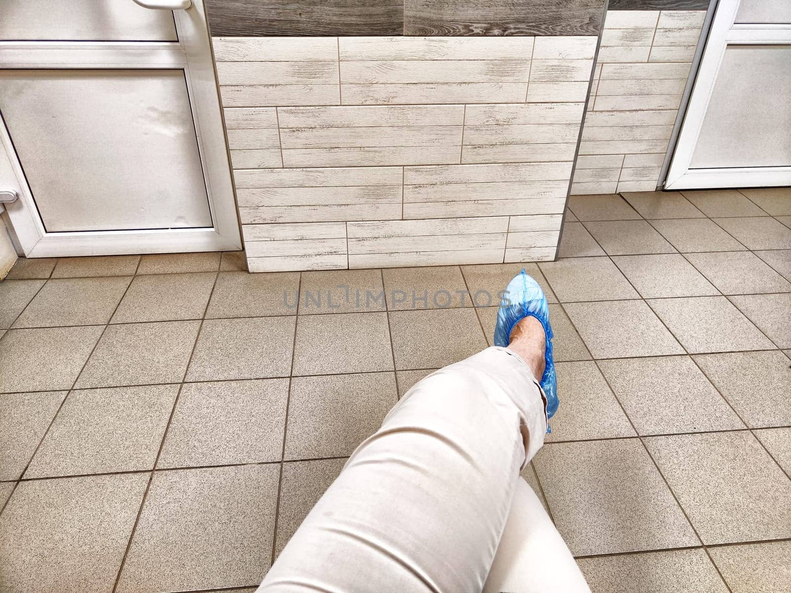 A person is waiting in a doctor's clinic hallway, sitting with feet in blue shoe covers on tiled flooring