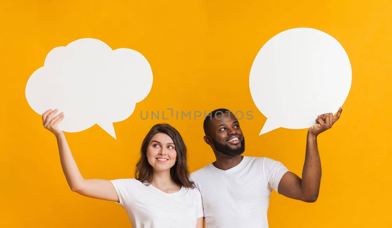 Happy multiracial couple looking at two empty communication bubbles above their heads and thinking about options, yellow background, panorama