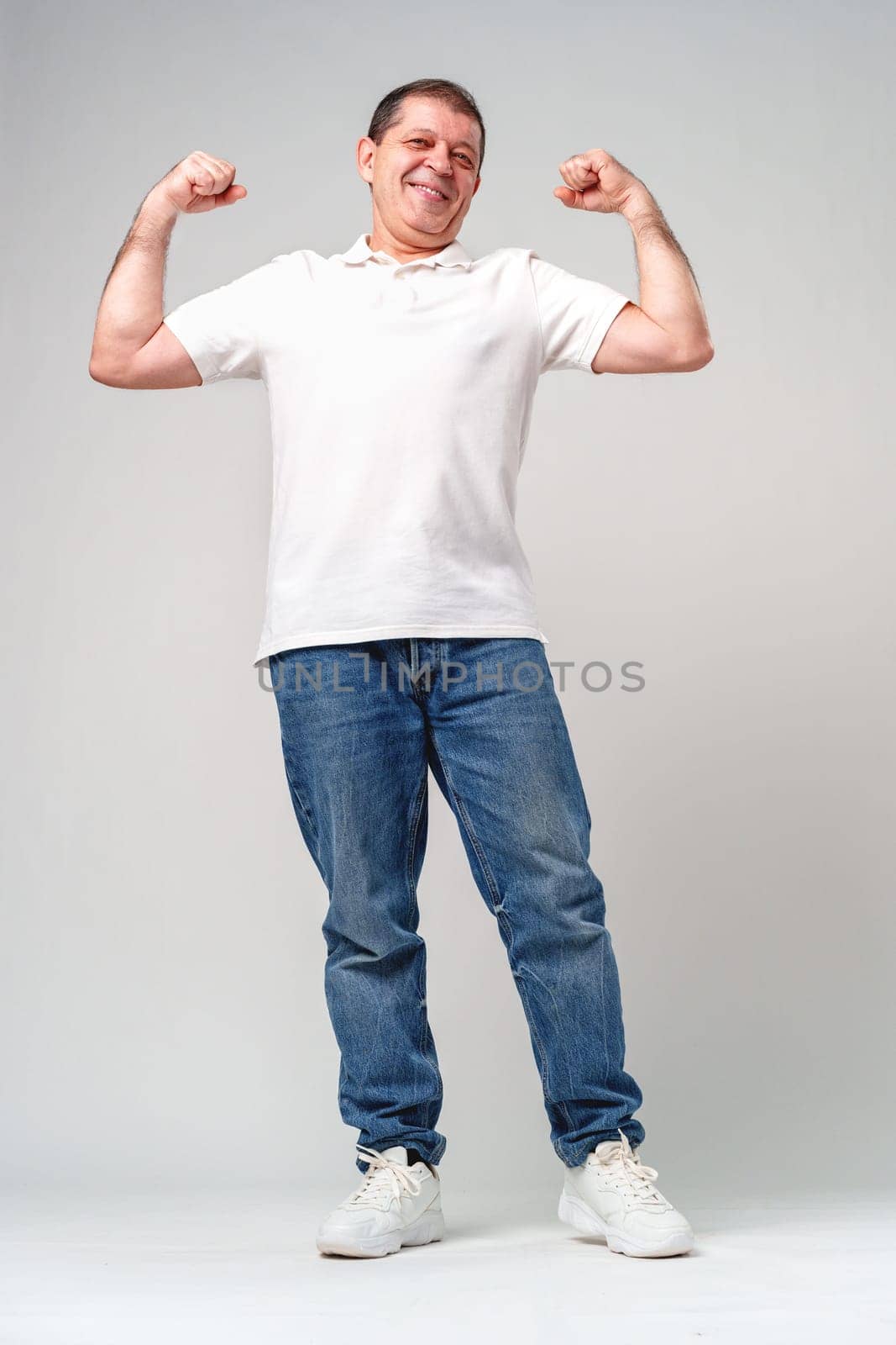 Happy older man showing off his strong biceps while standing against a light gray background close up