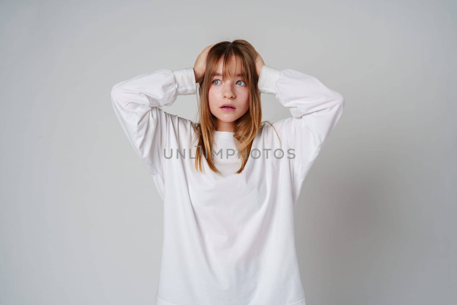 A teen girl in a loose white sweater looks surprised with her hands on her head against a plain gray backdrop close up