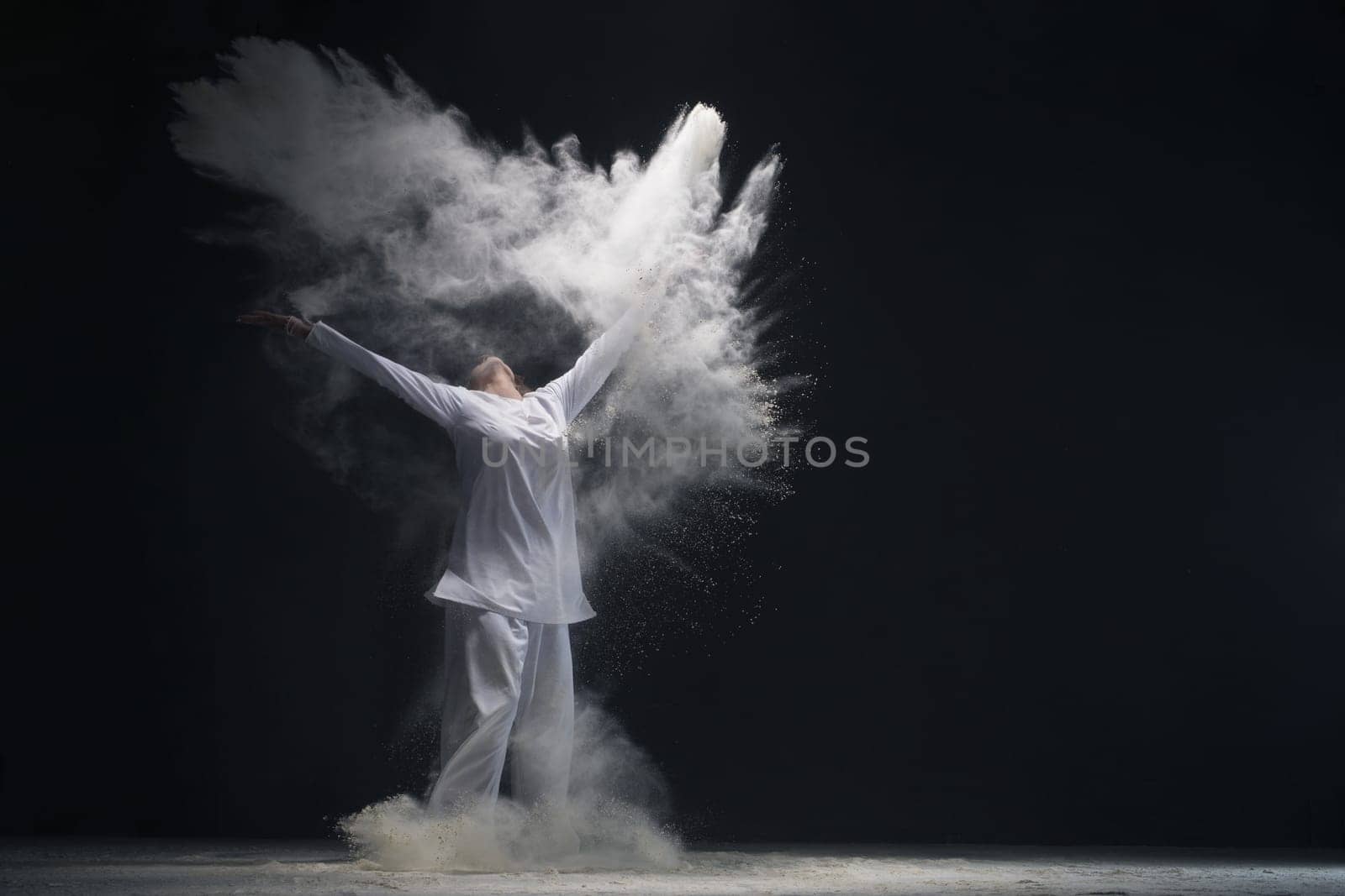 Man wearing white shirt and trousers jumping in white dust cloud in dark room full-length shot