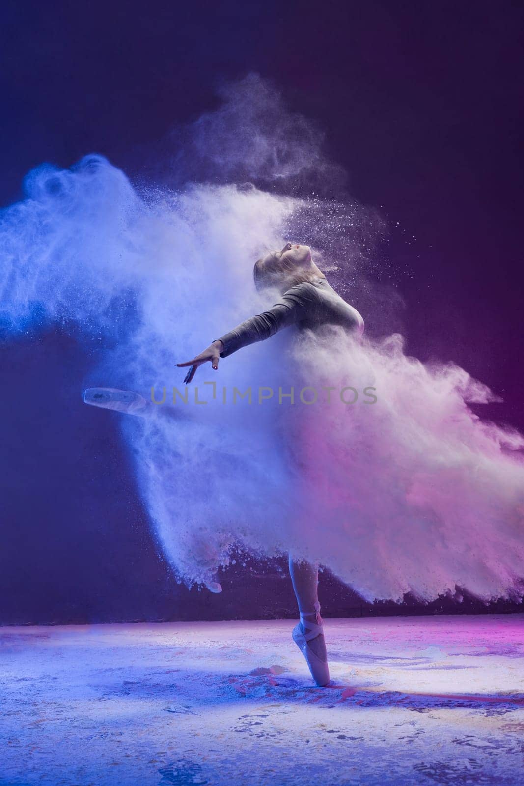 Woman in undrewear and pointe shoes jumping gracefully in a white dust cloud and color light profile view