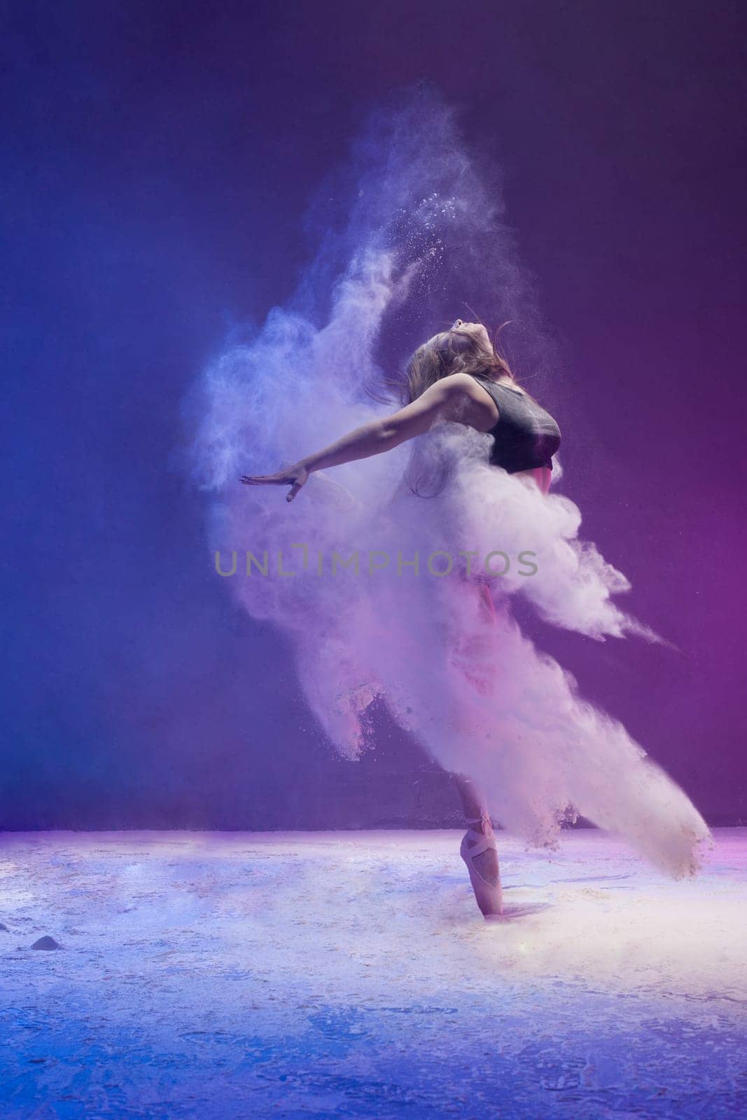 Woman in undrewear and pointe shoes jumping gracefully in a white dust cloud and color light profile view