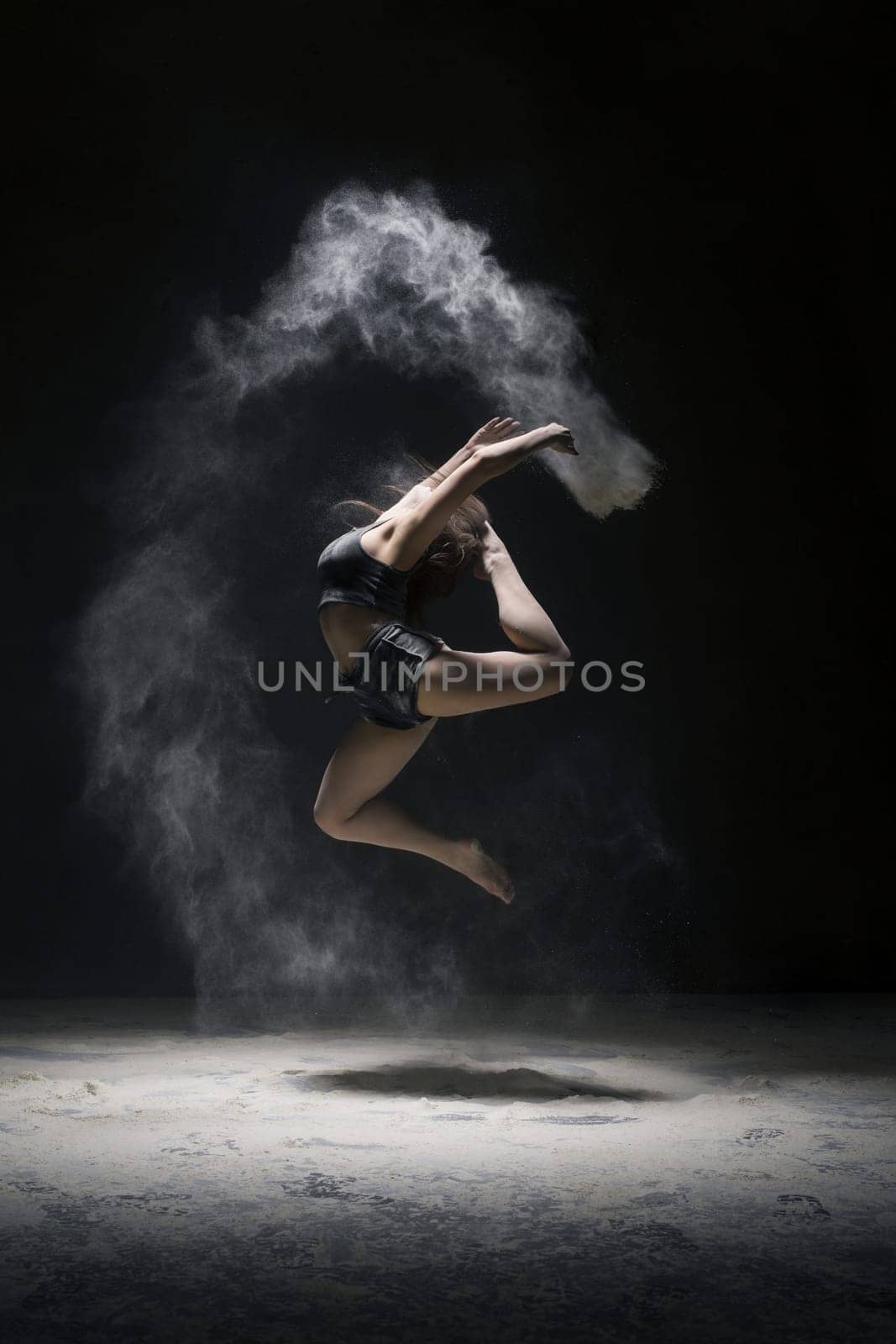 Woman in black shorts and top jumping gracefully in a white dust cloud profile shot in dark room