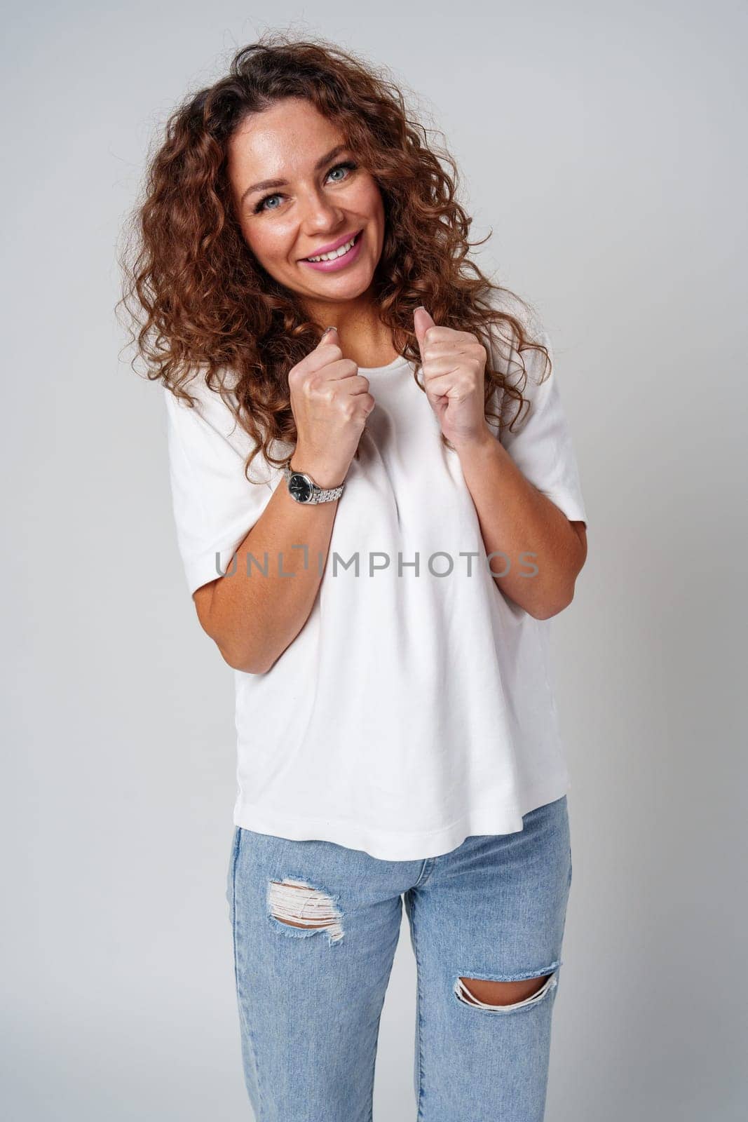 Cheerful woman with curly hair wearing a white shirt and jeans poses happily against a light gray background close up