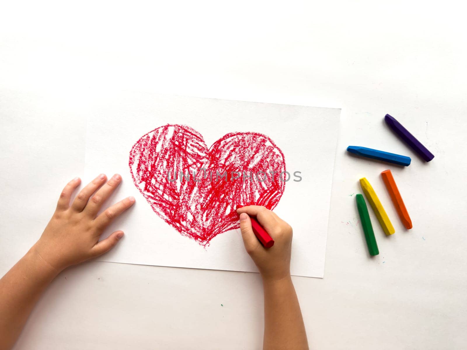 Child drawing a red heart with crayons on white paper.
