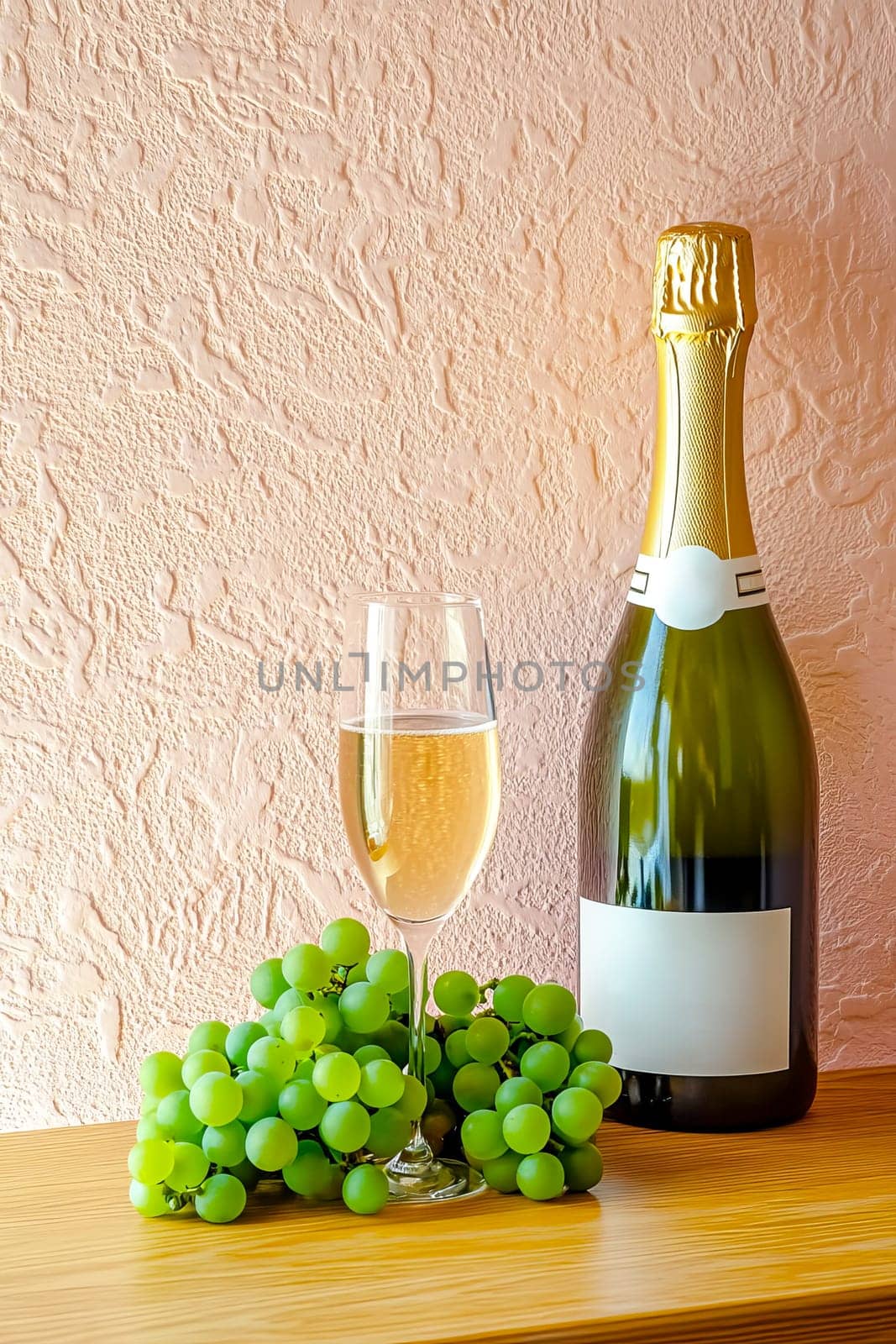 A bottle of champagne, a glass and a bunch of green grapes on a wooden table against a pink-gray textured plaster wall.