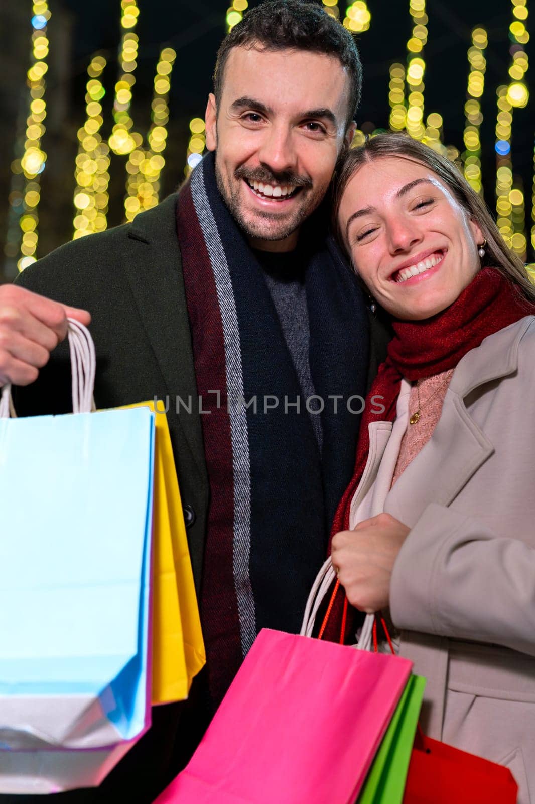 Smiling couple holding multiple colorful shopping bags, enjoying a festive evening out in the city. Dressed warmly for winter, they radiate joy and excitement, surrounded by holiday lights and a lively atmosphere.