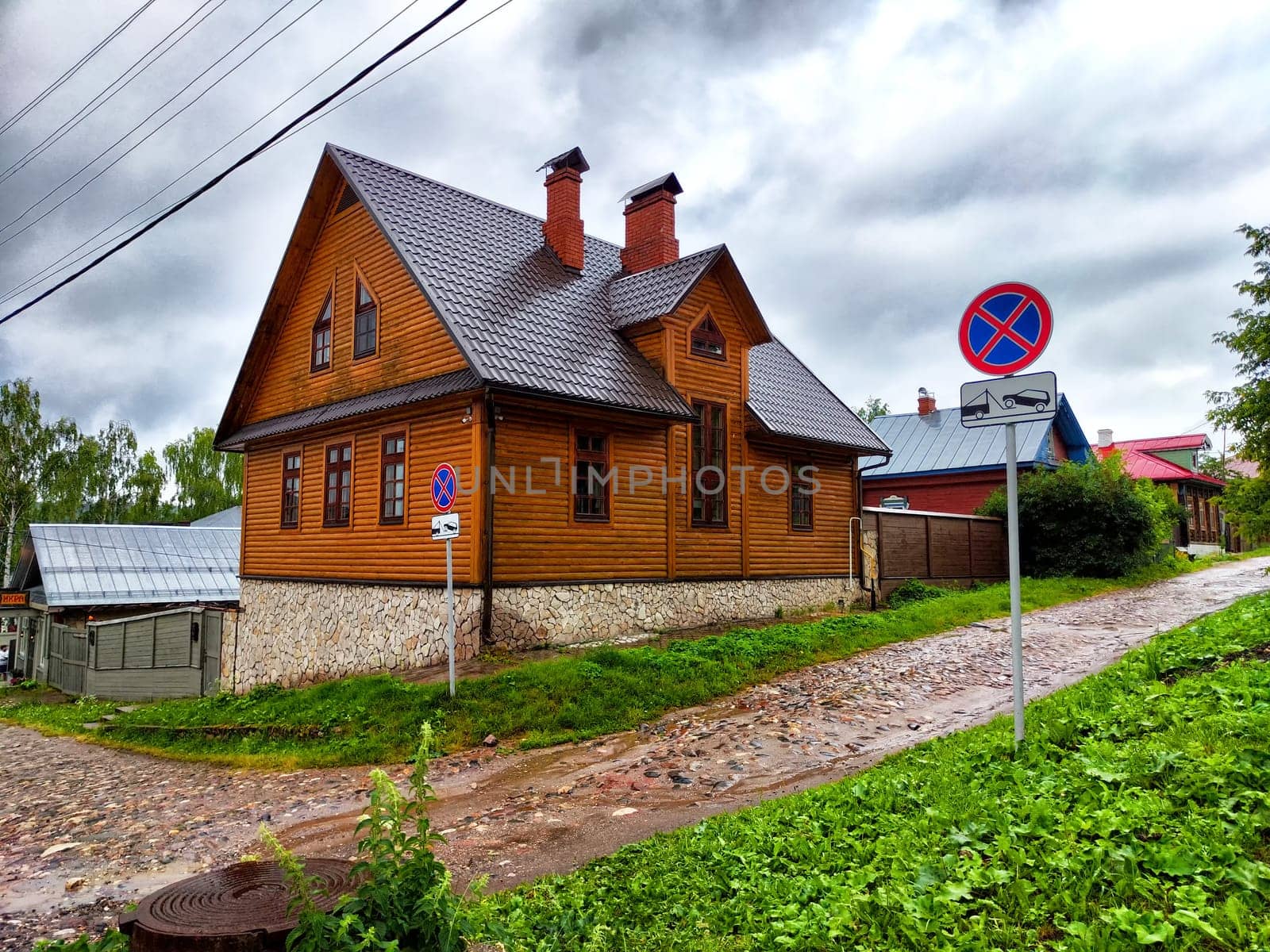 A charming wooden house stands on a hillside street, featuring rustic architecture and surrounded by lush greenery under gray clouds.