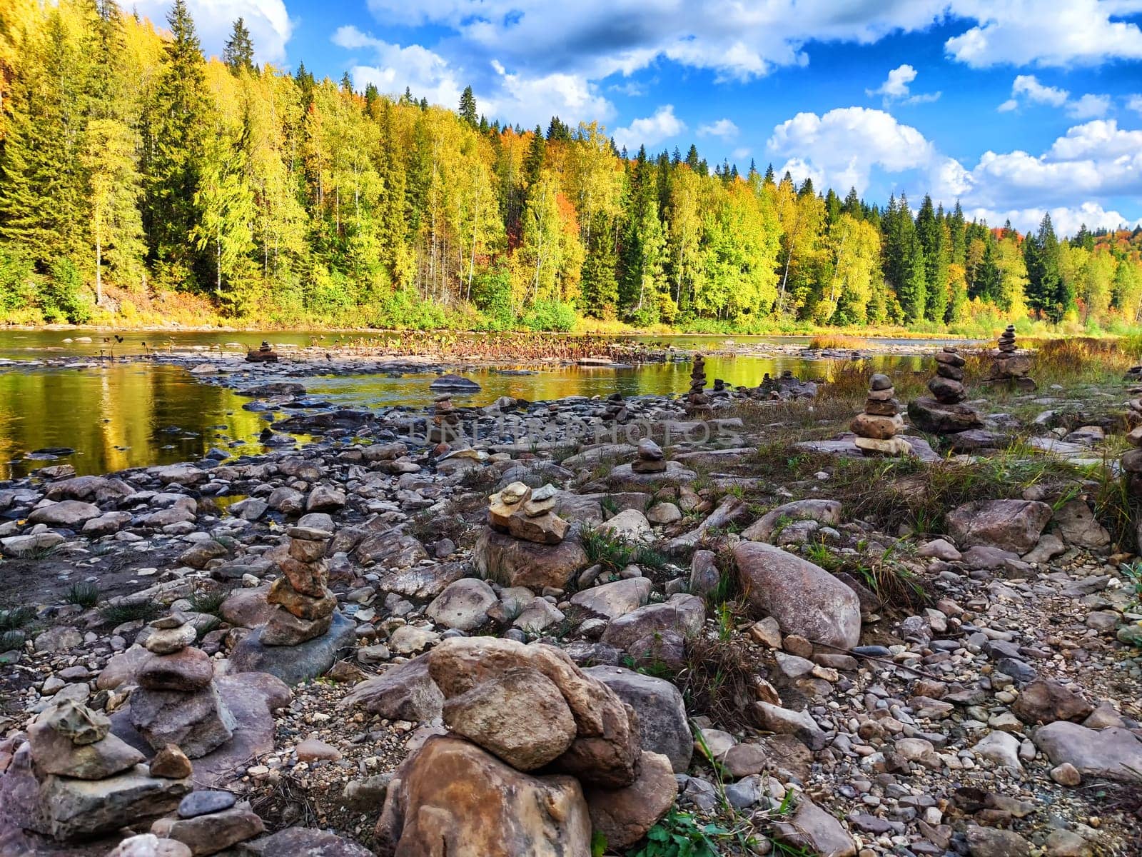 A calm riverbank features creatively stacked stones as colorful autumn foliage reflects serenity and nature\'s beauty.