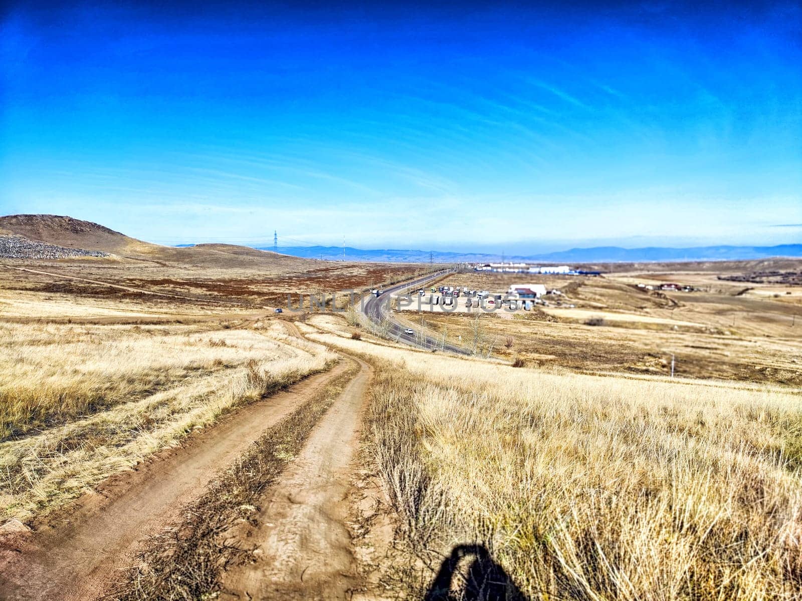 A dirt road meanders through dry grasslands, showcasing rugged hills and a small town in the background under a vibrant blue sky.