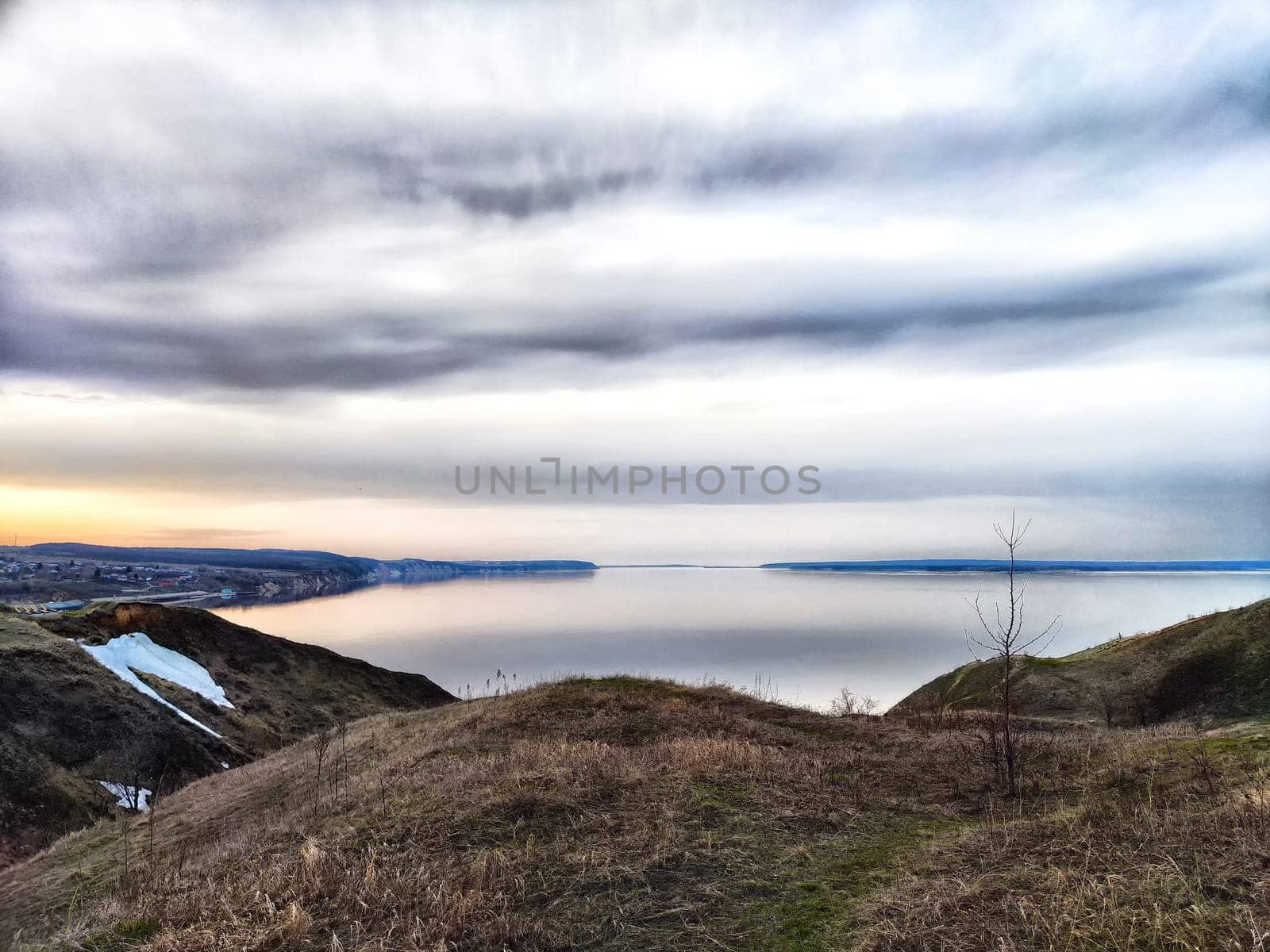 A serene river reflects soft pastel colors as the sun sets behind rolling hills, creating a tranquil atmosphere in early spring.