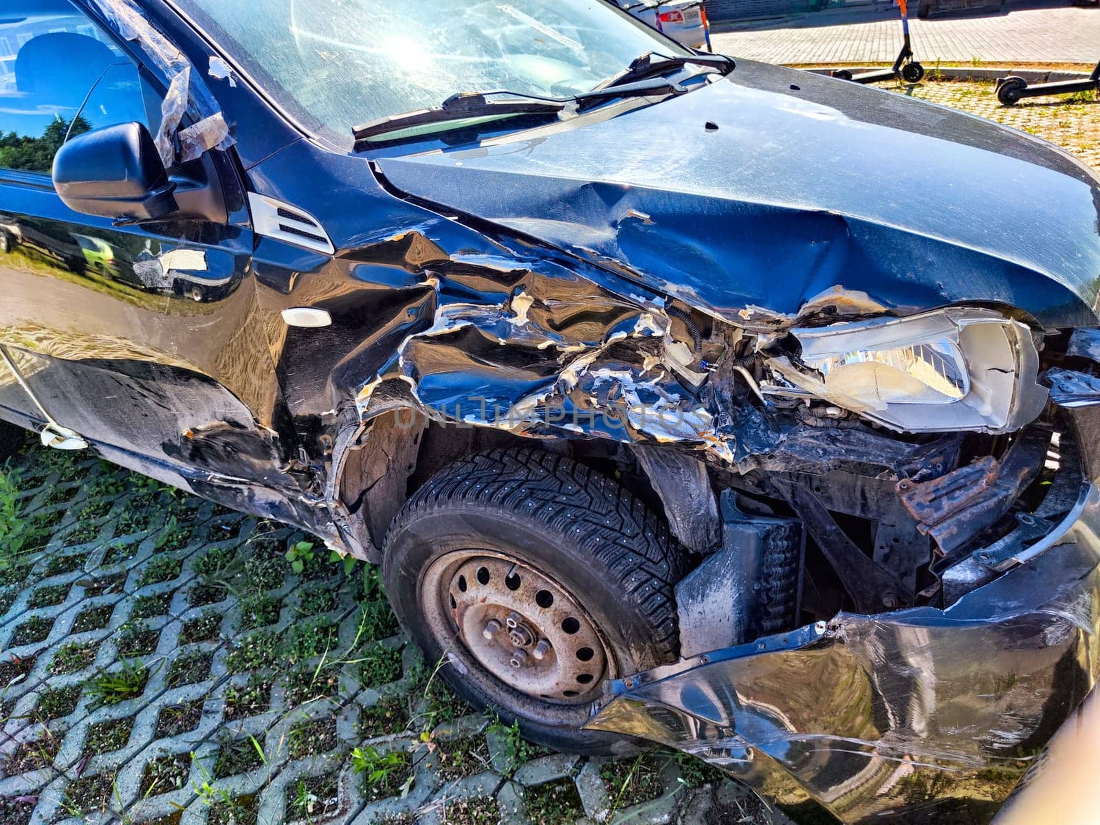 A black car shows significant damage to the front left side, parked near contemporary buildings in a sunny outdoor area