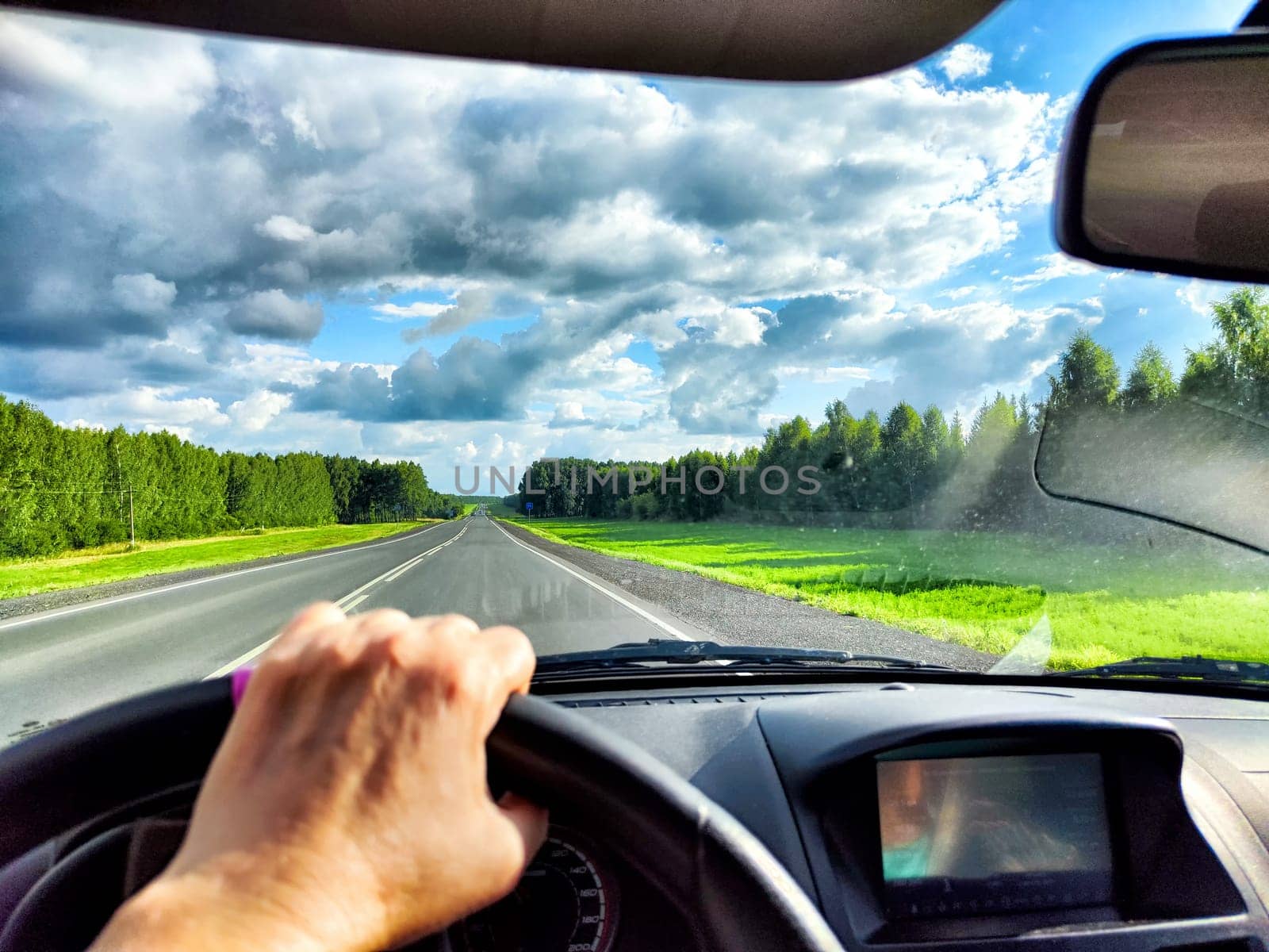 The driver steers along highway, with vibrant green fields and dramatic clouds stretching across the sky