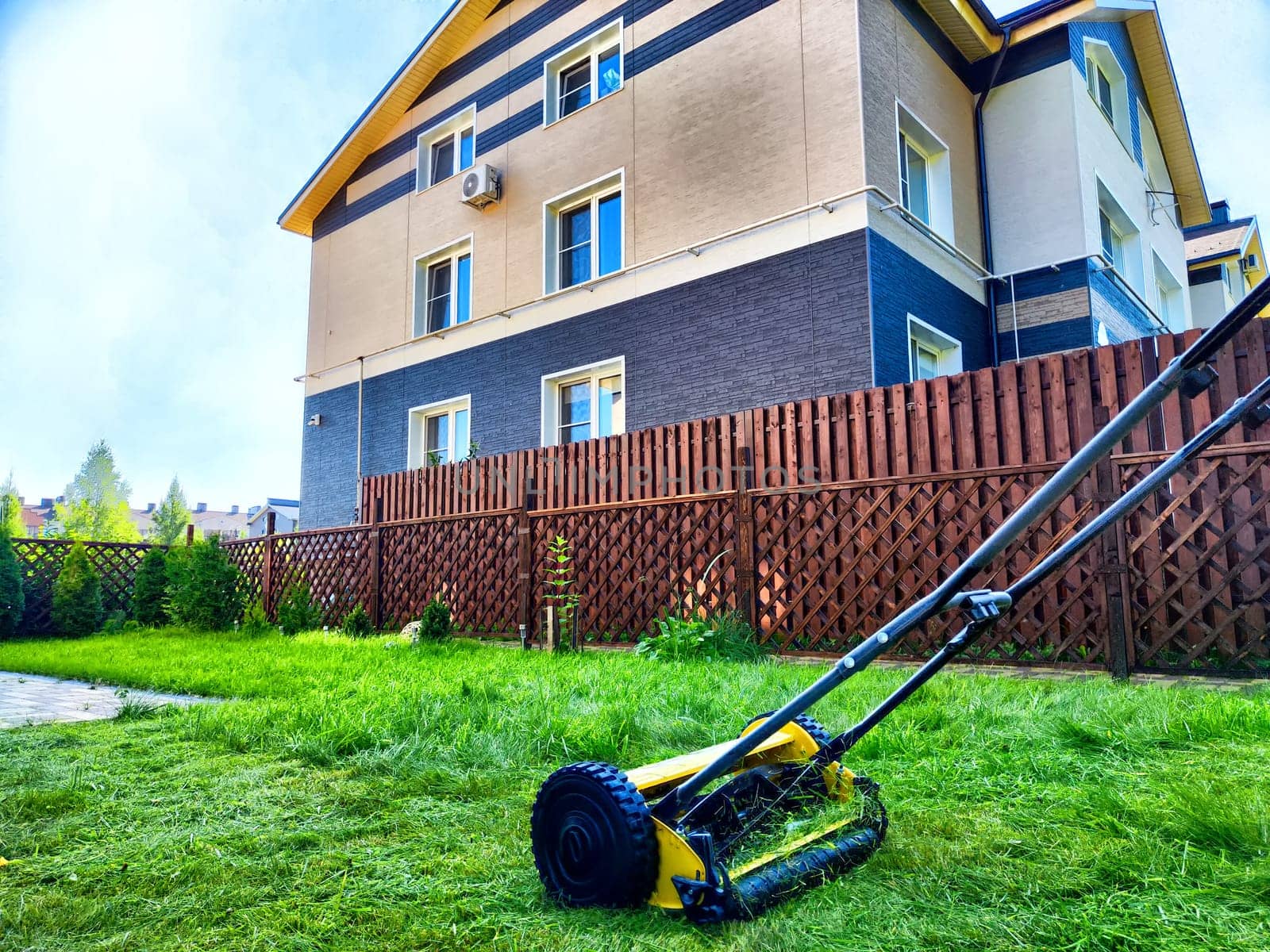 A lawn mower is actively trimming the green grass in a tidy yard next to a contemporary house during a sunny day.