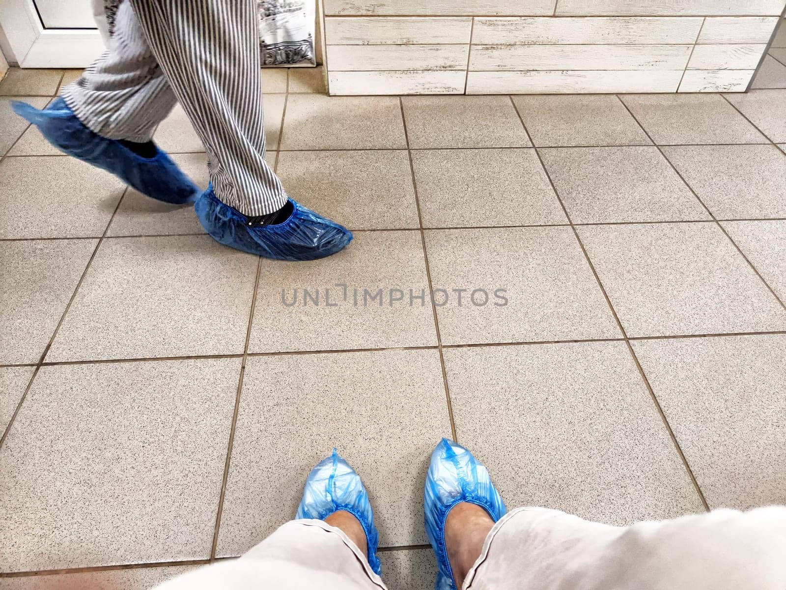 A person is waiting in a doctor's clinic hallway, sitting with feet in blue shoe covers on tiled flooring