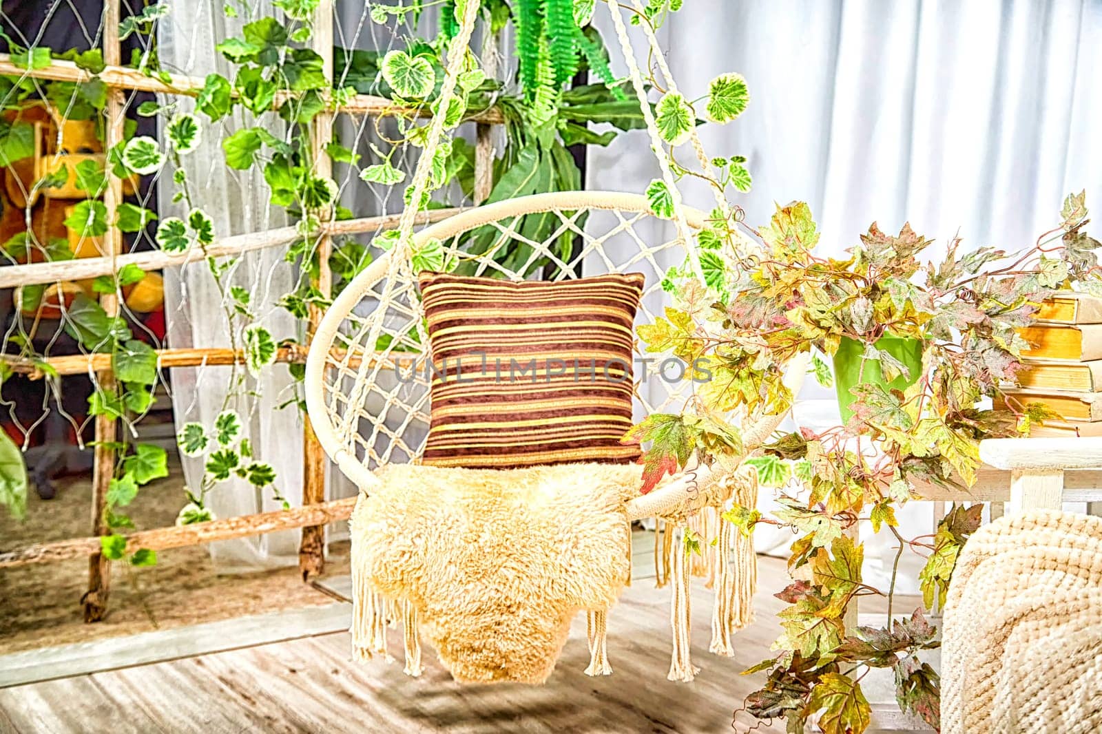 Modern cozy beautiful room with a braided rope macrame swing, chair, green plants, small table and curtains. Interior and background. Location for photo shooting