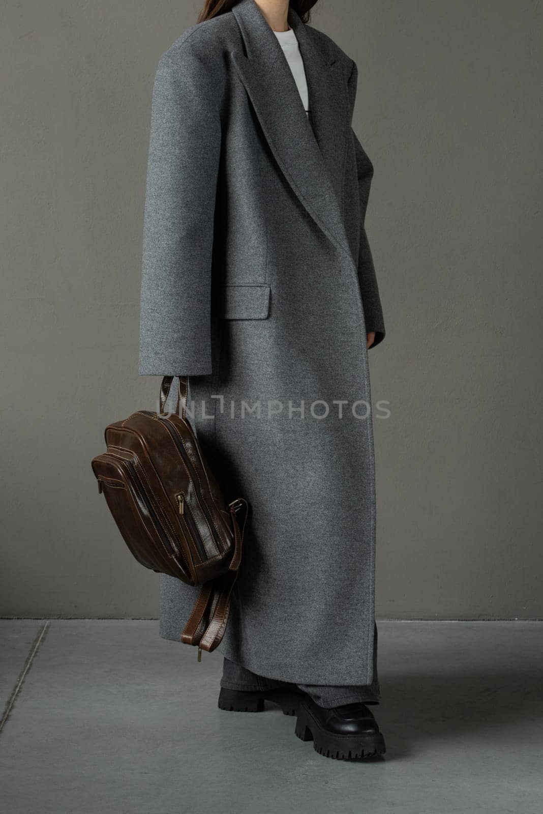 woman posing with a leather backpack wearing a warm gray coat and black boots.