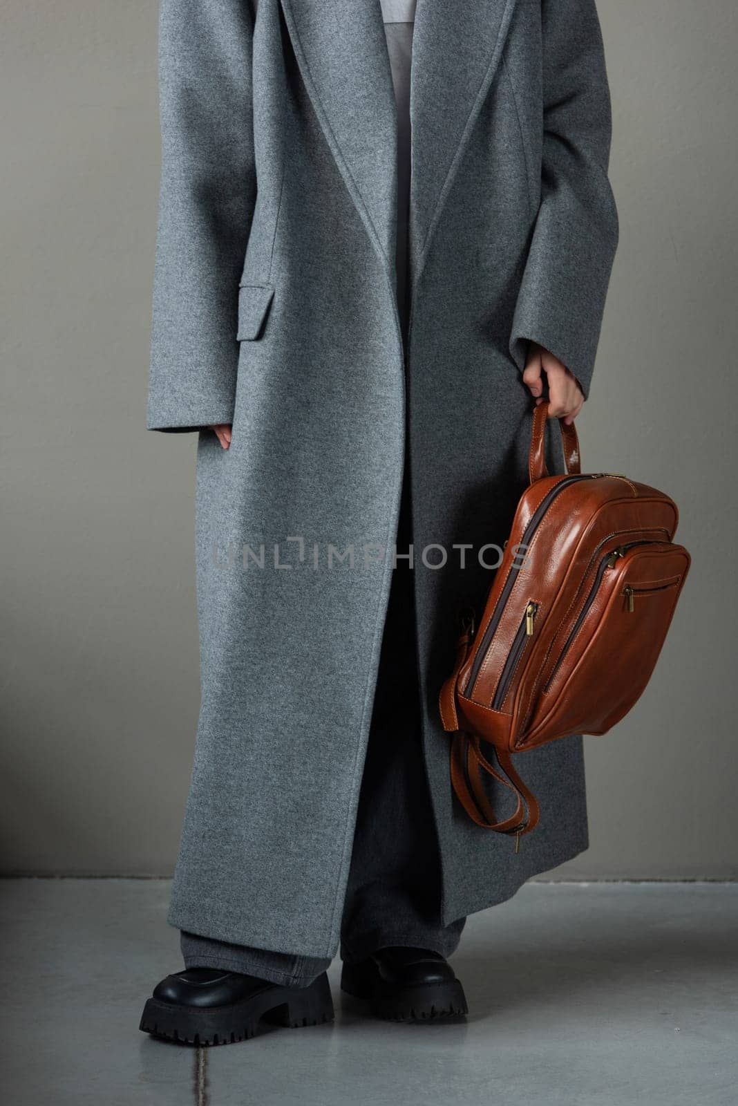 woman posing with a leather backpack wearing a warm gray coat and black boots.