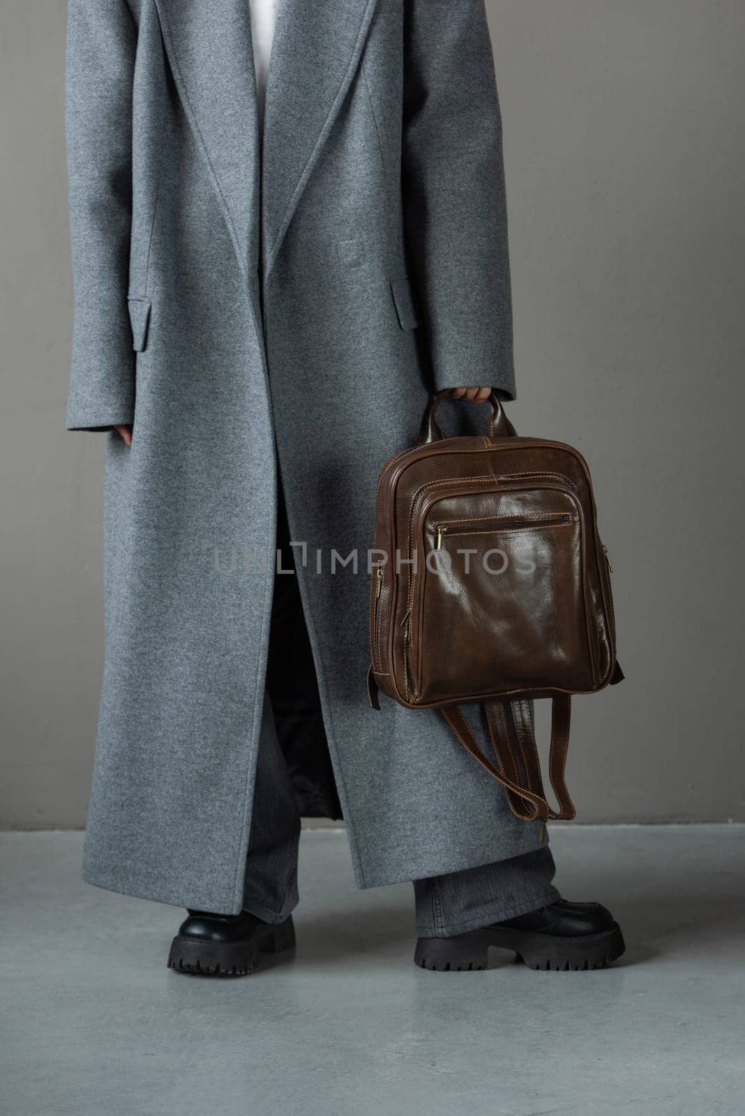 woman posing with a leather backpack wearing a warm gray coat and black boots.