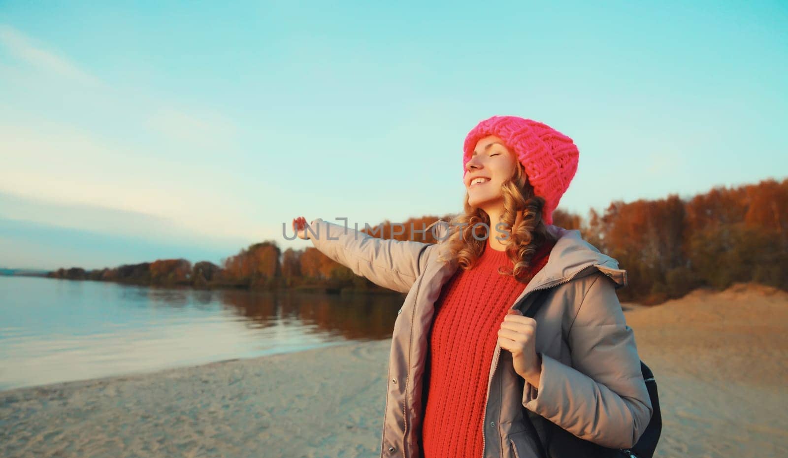 Travel, adventure, happy traveler young woman on the beach with beautiful sunset, enjoying sunny landscape, warm weather, standing on sea coast, wear backpack, hat, jacket, nature background