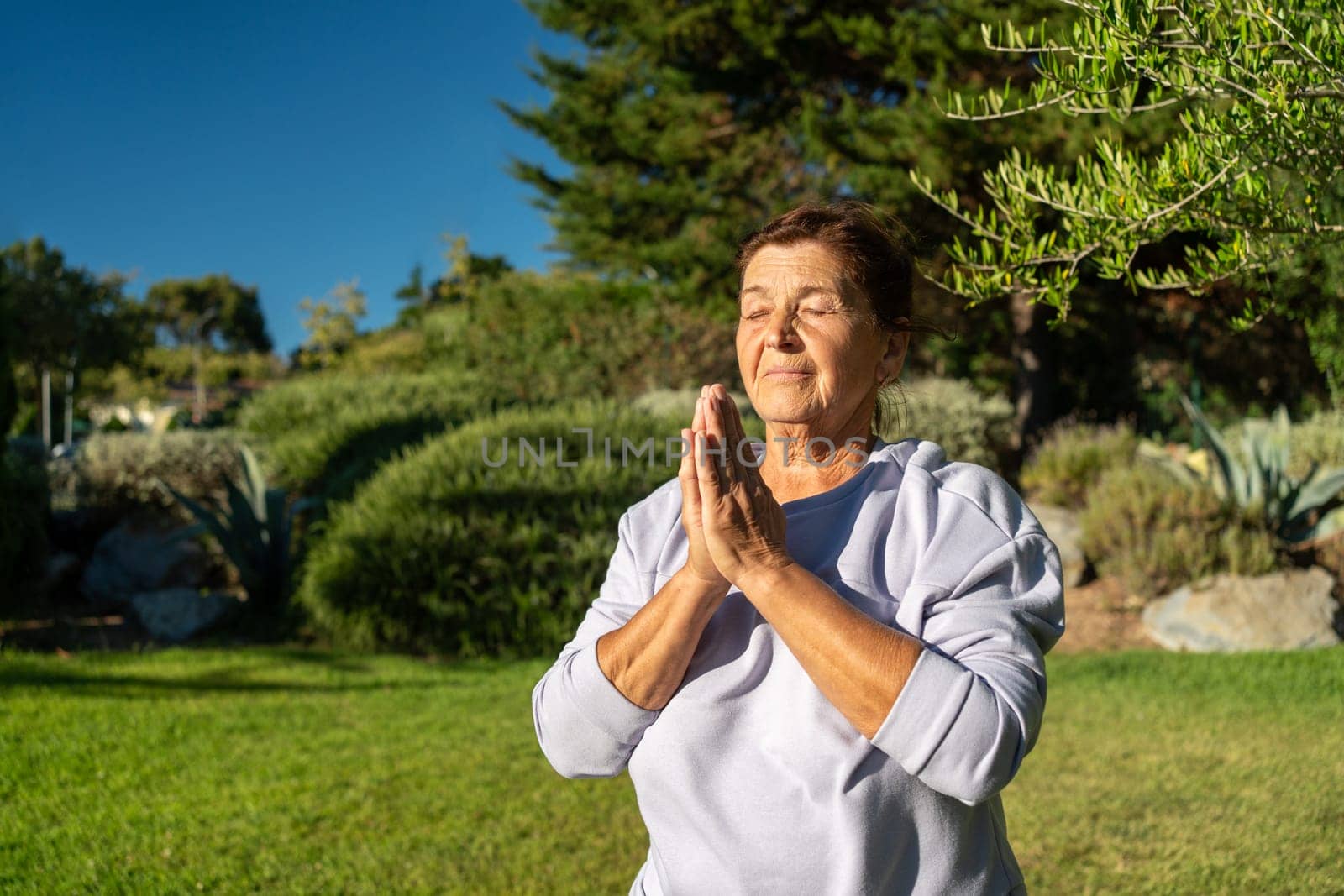 Smiling senior woman in gray sportswear practicing prayer pose in bright park. She stands with hands clasped in yoga posture during daytime
