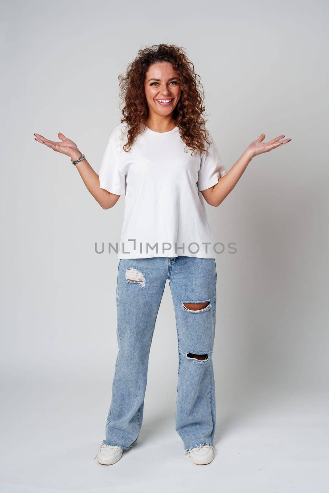 Cheerful woman with curly hair poses with open arms in a white shirt against a plain background close up